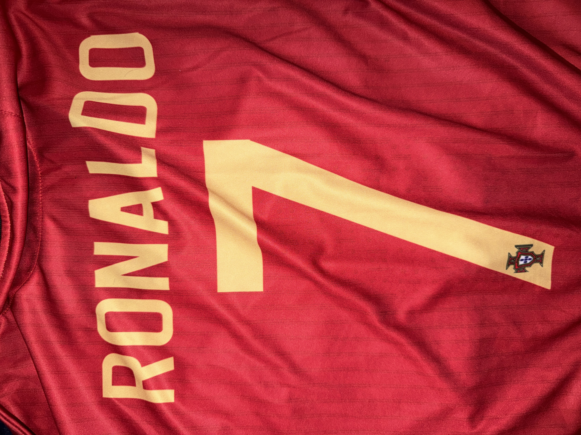 The image shows a close-up, top-down view of a red soccer jersey with "RONALDO" printed in gold block letters across the top, followed by a large gold number "7" below it. A small crest, the Portuguese coat of arms, is visible at the bottom left corner of the number 7. The jersey is made of a textured fabric with subtle vertical ribbing and is creased in several places due to being laid flat. There are no people, interactions, or environmental details visible, and the location in Nikopol, Bulgaria, is not indicated in the image itself.