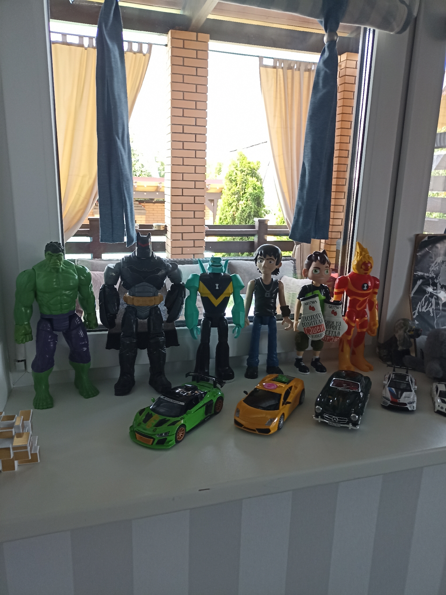 In an indoor setting with natural light, several action figures and toy cars are displayed on a white ledge. The figures, arranged from left to right, include a green Hulk figure, a black Batman figure, a turquoise and black robot figure, two humanoid figures, and a red and yellow humanoid figure. In front of the figures are several toy cars: a green sports car, a yellow sports car, a black classic car, and a white and blue race car. The background shows a window with beige curtains and a brick pillar structure, suggesting an outdoor patio or balcony visible through the window. The overall scene is a collection of toys on display.