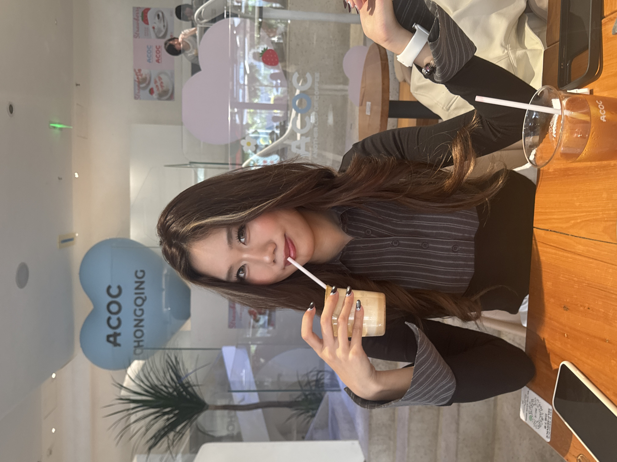 A woman is seated at a wooden table, holding a glass of a light-colored beverage with a straw and drinking from it. She is wearing a dark, striped button-down shirt and black pants. Her long hair is dark with light streaks, and her fingernails are painted. On the table, next to the glass, is a black smartphone.
Behind the woman, a glass wall is visible, revealing the interior of a business named "ACOC" which appears to be located in 江北城街道, China. A heart-shaped balloon with "ACOC CHONGQING" printed on it can be seen in the background. A plant is also visible in front of the glass wall. Another person is partially visible in the background.