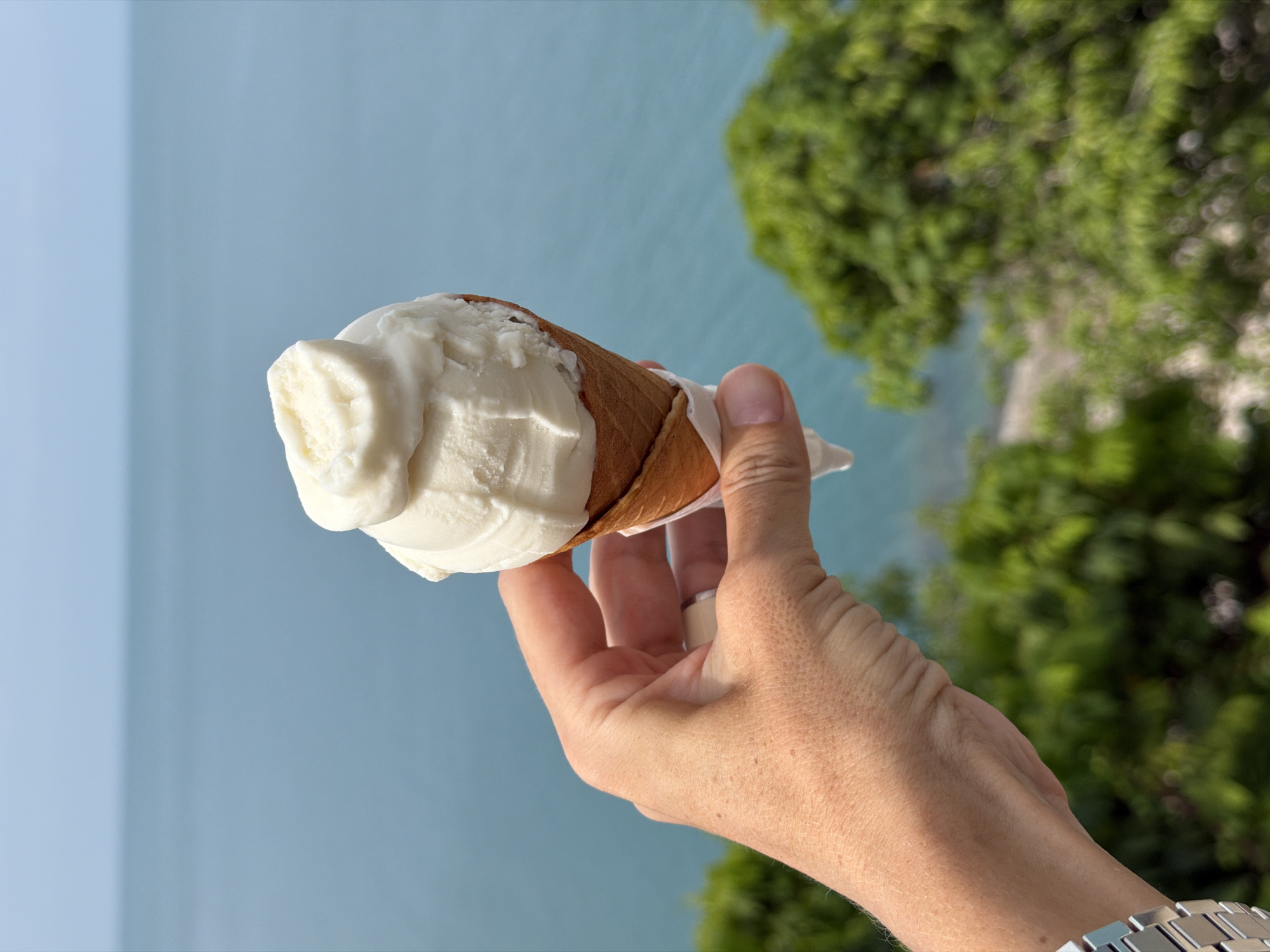 In an outdoor setting with a blurred ocean and lush green foliage in the background, a person's hand is holding a vanilla ice cream cone. The ice cream is white and creamy, with a slightly uneven top. The cone is a light brown waffle cone, partially wrapped in white paper. The hand is light-skinned, with a silver watch on the wrist and a ring on one finger. The scene appears to be a scenic overlook in Ko Samui District, Thailand, given the tropical vegetation and the ocean view.