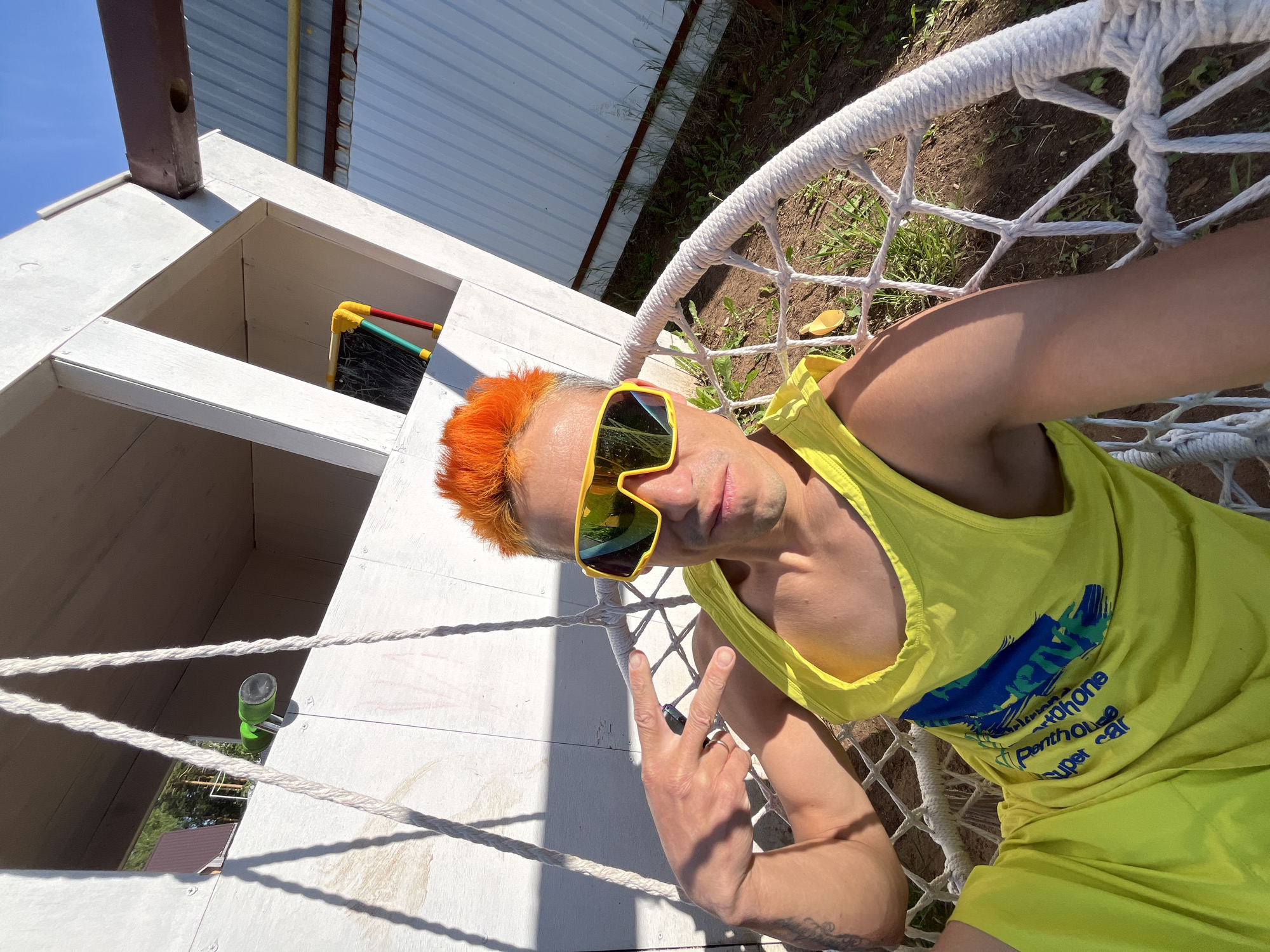 An adult male with bright orange spiky hair and large yellow mirrored sunglasses is seated in a white woven rope swing chair outdoors in Chaikovsky, Russia. He wears a neon yellow tank top with a blue and green graphic print that includes partial text such as "Penthouse" and "super car," and matching neon yellow shorts. His left arm is raised, with his hand forming a peace sign (V-sign); a silver-colored ring is visible on his left ring finger, and tattoos are present on his left arm. The background features a white wooden or plywood structure, possibly a playhouse or shed, with visible screw fasteners. Mounted on the left side of this structure is a green cylindrical object with a black end. On the right side, within an opening of the structure, a small, colorful framed object with yellow, red, and green sections and a dark surface is visible. Beyond the immediate structure, a brown roof and a corrugated metal wall are discernible, along with sparse green vegetation and bare soil on the ground. A bright blue sky is visible above, and strong shadows indicate direct sunlight.