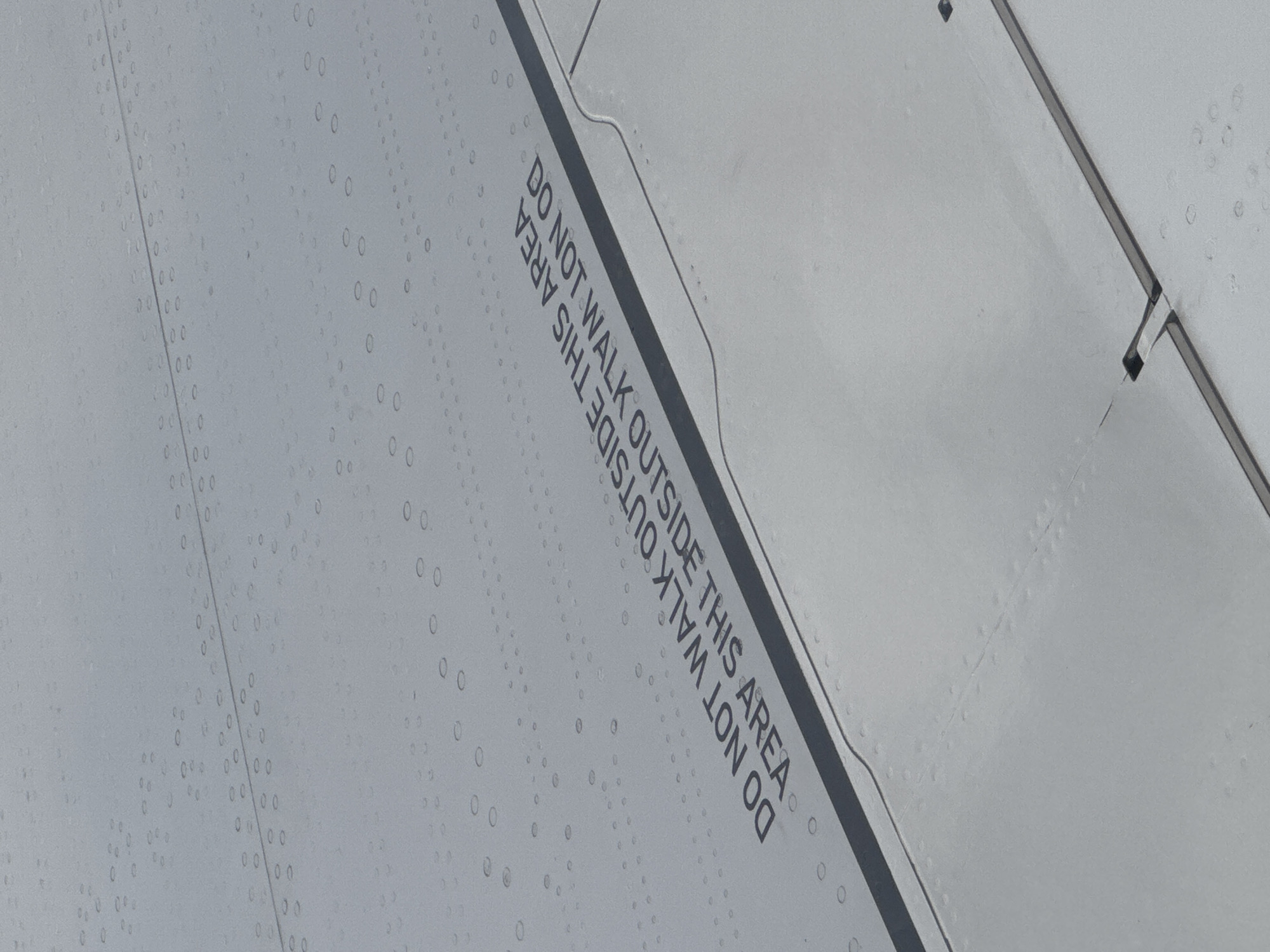 The image shows a close-up of an airplane wing, likely a passenger aircraft, with rivet lines and a clearly marked "DO NOT WALK OUTSIDE THIS AREA" warning. The wing's surface is clean and gray, with what appears to be condensation or light droplets present. The wing appears metallic in color. There is no evidence to determine the specific location of the photo.