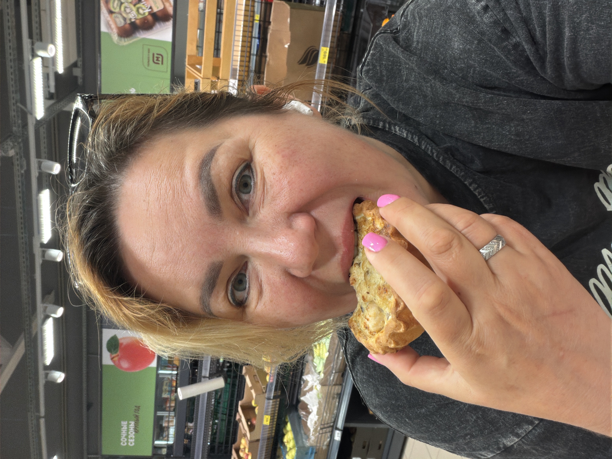A woman is holding a pastry in her right hand and taking a bite. She is wearing a dark gray denim jacket, a ring on her ring finger and has pink nail polish. She is in what appears to be a grocery store. The background includes shelving with various products. The environment suggests a retail or shopping location. Signs in the background written in Russian suggest this location is in Russia. Further research indicates "Ленинградская" translates to "Leningradskaya," a place in Russia.