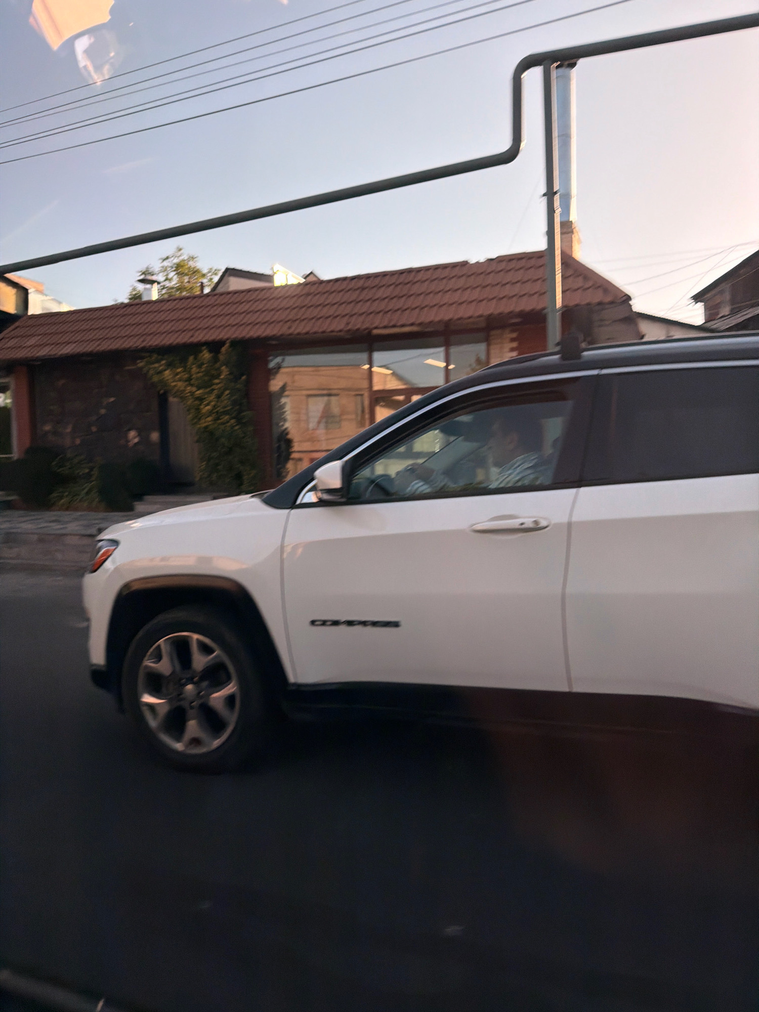 In Yerevan, Armenia, a white Jeep Compass is captured in motion, its side profile clearly visible. The vehicle is traveling on a paved road, with a building featuring a brown tiled roof and stone facade in the background. A person is visible driving the Jeep. Overhead, electrical wires and a metal pipe structure cross the frame, indicating an urban setting. The sky is clear and blue, suggesting daytime. The image has a slightly reflective quality, possibly from the car's windows.