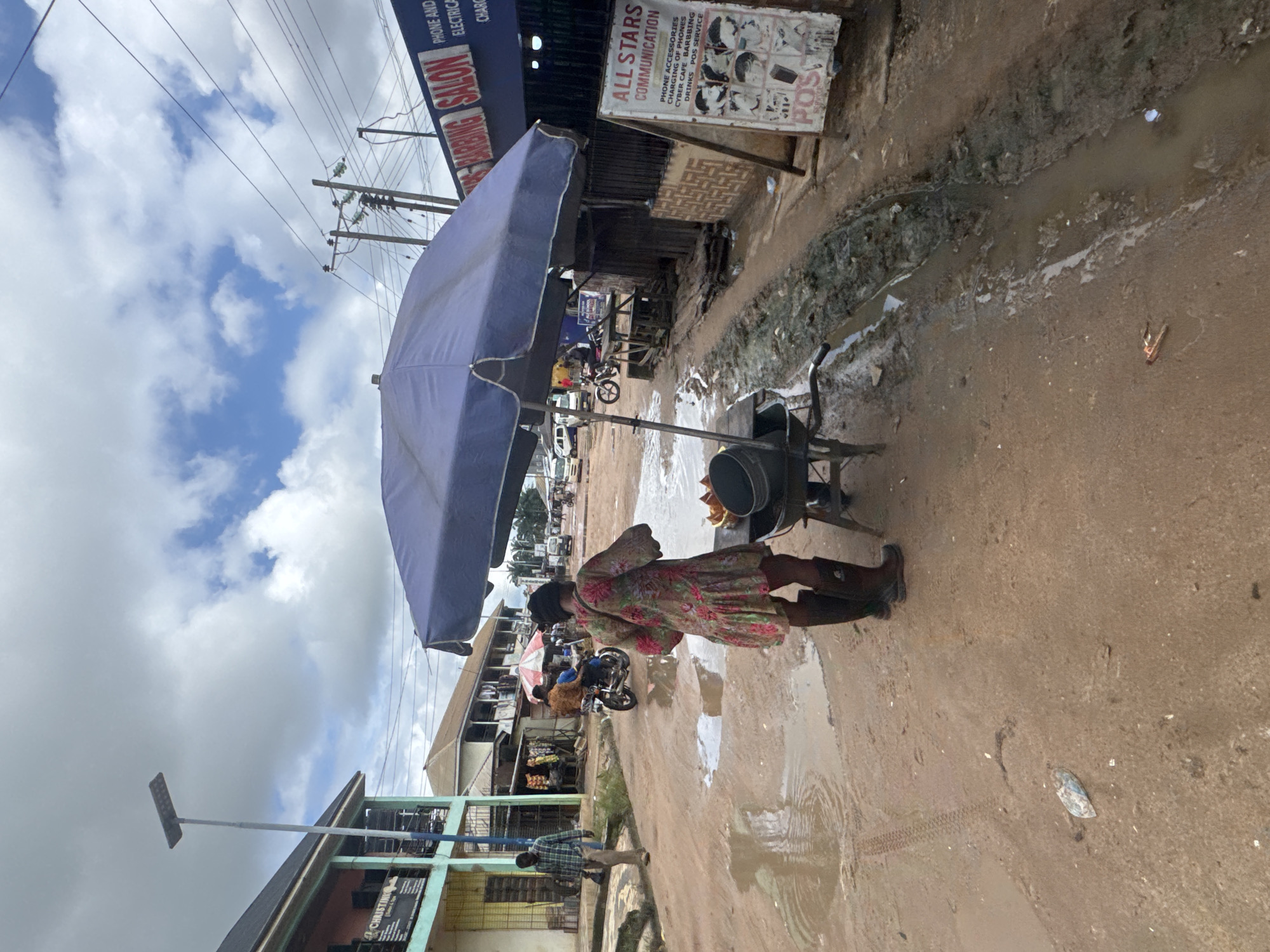 The image depicts a street scene in Ihiagwa, Nigeria, after a rainfall, characterized by muddy terrain and standing water. A woman, seen from behind, stands near a cart with a large blue umbrella, suggesting a vendor. She is wearing a floral dress and tall black boots. A motorcycle with two passengers is visible in the mid-ground, navigating the wet road. In the background, buildings line the street, some with shops, including one with signage for "Phone and Electrical Services" and another for "All Stars Communication." The overall environment appears to be an informal commercial area.