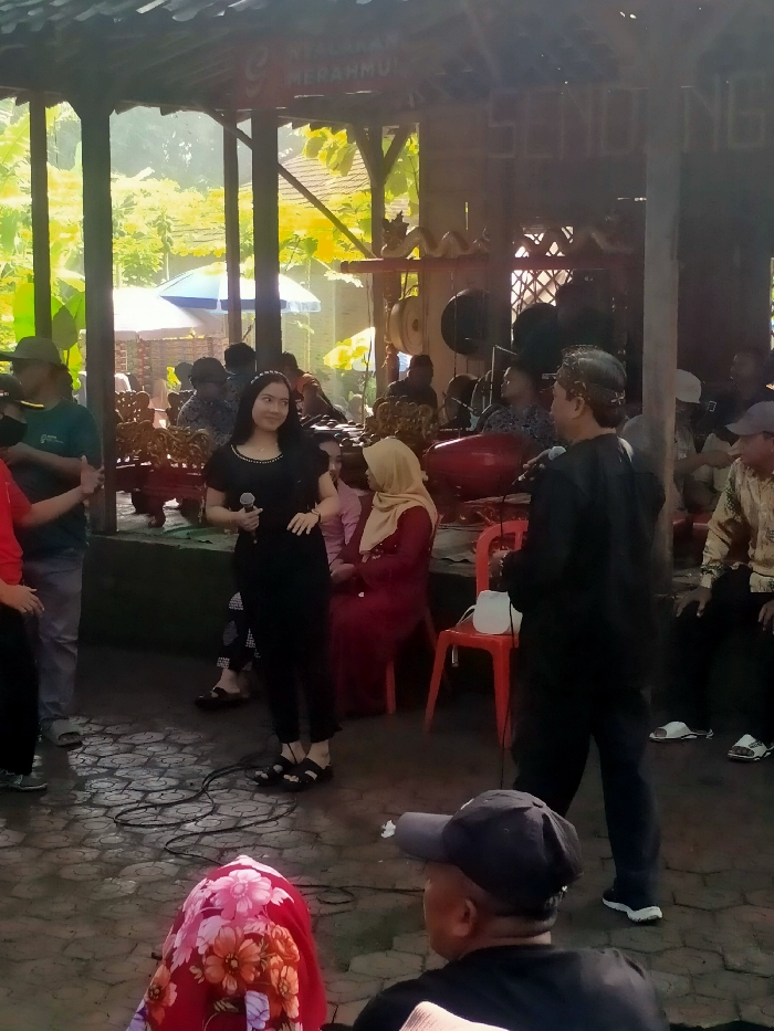 The scene takes place in a covered outdoor area with a tiled floor, under a wooden roof structure, in Masaran, Indonesia (latitude: -7.471, longitude: 110.957). A woman wearing a black dress and a microphone is standing in the center, facing the right side of the image. A man, wearing a black jacket and a white hat, stands behind her, holding a microphone and facing the woman. To the right, a woman wearing a red hijab and a long red dress sits on a red chair, looking towards the woman with the microphone. Several other people are present in the background, some sitting on chairs, some standing, and some seated on a wooden structure to the left side of the scene. There are numerous decorative metal plates hanging from the roof structure and a red wooden structure with large red drums sits in the background behind the woman in the black dress. The wooden structure and the drums suggest that this is some sort of musical performance or event.