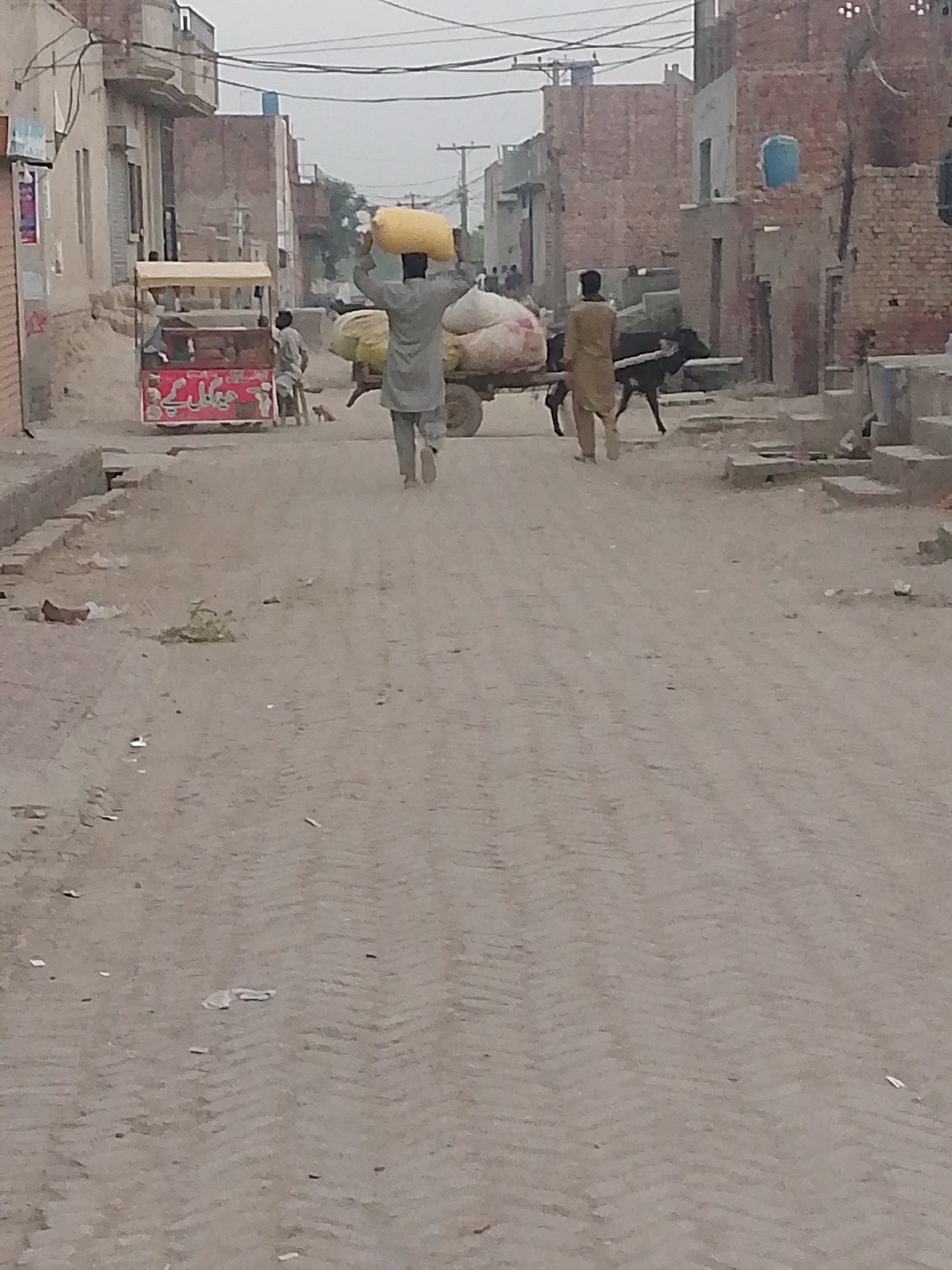 A man carrying a yellow bag on his head walks on a dirt road in a rural area. Another man follows behind pulling a cart with two large bags. A black cow pulls the cart. In the background, a food cart is seen next to a man sitting on a stool. Buildings are visible on both sides of the road.