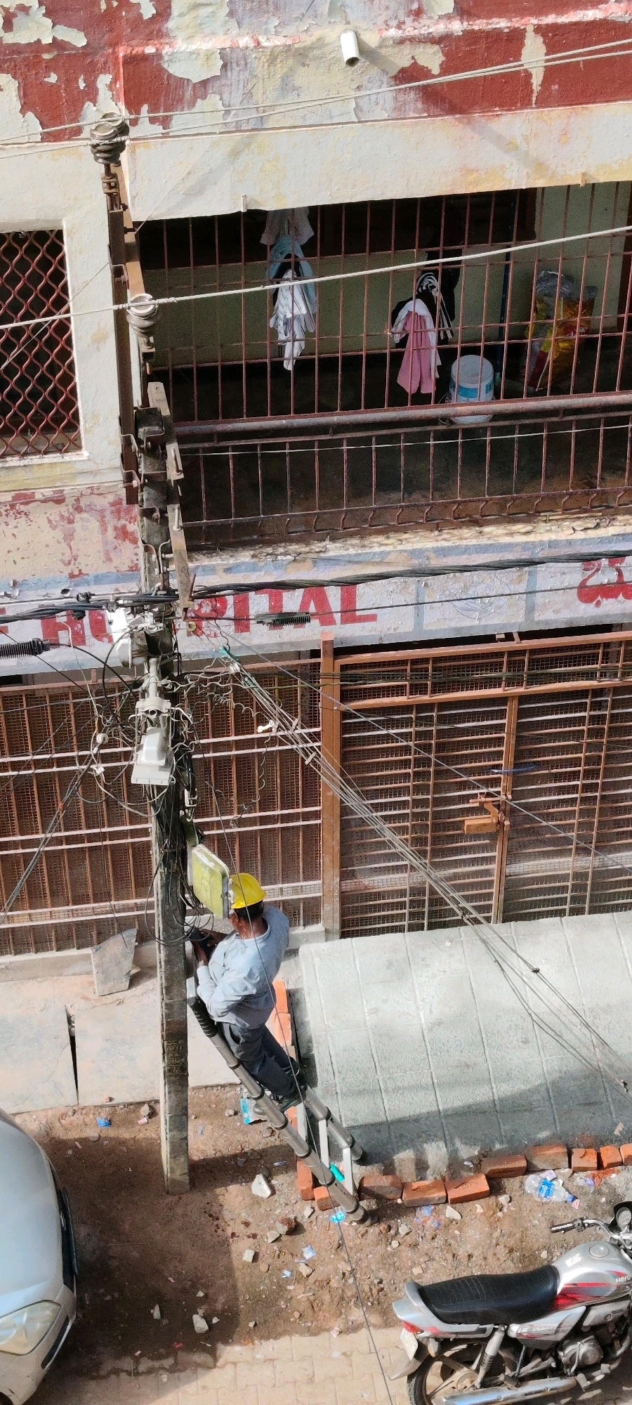 An overhead, high-angle, medium shot shows a man in a yellow hard hat and a light blue shirt working on a utility pole in a street in Belagavi, India. The man is on a ladder, and his hands appear to be holding some tools as he works on the numerous wires and cables attached to the pole. The utility pole is heavily laden with wires, transformers, and other electrical equipment. The man is positioned on the pole, approximately halfway up.
Behind the man and the pole is the facade of a building with an upper story featuring barred windows and laundry hanging on a line. Below this, a sign with red lettering that reads "OSPITAL" is visible on the building's facade, suggesting the building is a hospital. The street level shows a silver car on the left and a silver motorcycle on the right. The ground is a mix of dirt, gravel, and scattered debris, including bricks and plastic bottles. The overall impression is of a busy urban street with ongoing utility work.