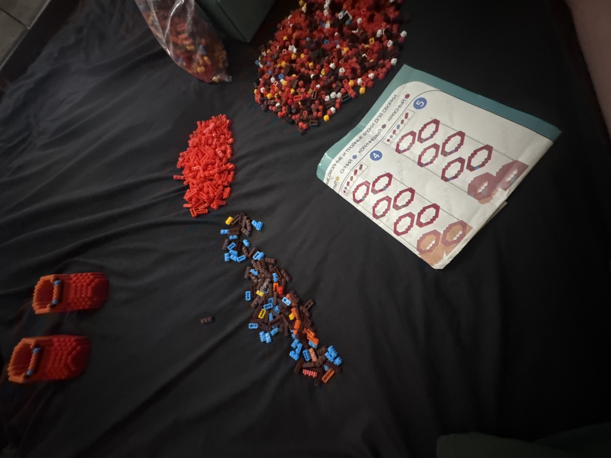 The image displays a top-down view of a dark, textured surface, possibly a bedsheet or fabric, with various construction toy pieces scattered across it. Two red objects resembling small boots or containers are positioned in the upper left quadrant. A pile of red toy bricks is located in the upper right of the image, with a clear plastic bag filled with red, yellow, blue, and brown toy pieces to its right. A cluster of blue, brown, and red toy bricks is spread in a diagonal line from the center-left towards the top. In the bottom right quadrant, an instruction booklet is open, showing diagrams of toy construction with numbered steps and color indicators, likely for building. The scene is an indoor environment, and there is no indication of the city of Makiivka, Ukraine.