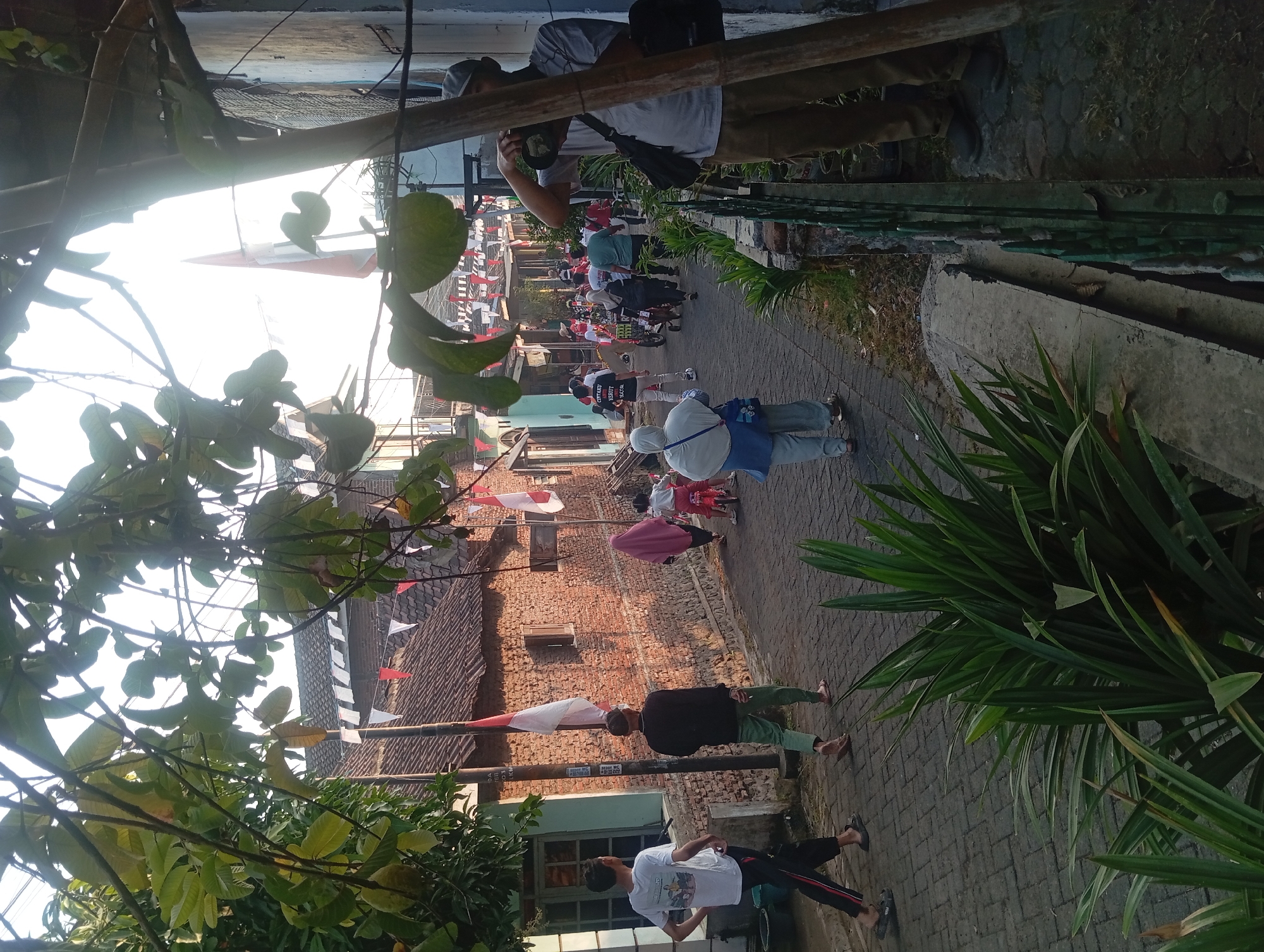 In Jogosari, Indonesia, a narrow, cobblestone alleyway is lined with brick buildings and adorned with red and white pennant flags, indicating a celebration. In the foreground, lush green plants obscure the lower portion of the frame. Several individuals are visible walking or participating in an event. On the right, a man wearing a gray t-shirt and brown pants is taking a photograph with a camera. Further down the alley, a young man in a white t-shirt and black pants walks to the left, and a person in a black shirt and green pants walks to the right. A woman in a pink hijab and a child on a red tricycle are also present. In the background, a parade or procession is underway, with more people walking and what appears to be a cart or small vehicle. The sun is low, casting long shadows and illuminating the scene with warm light.