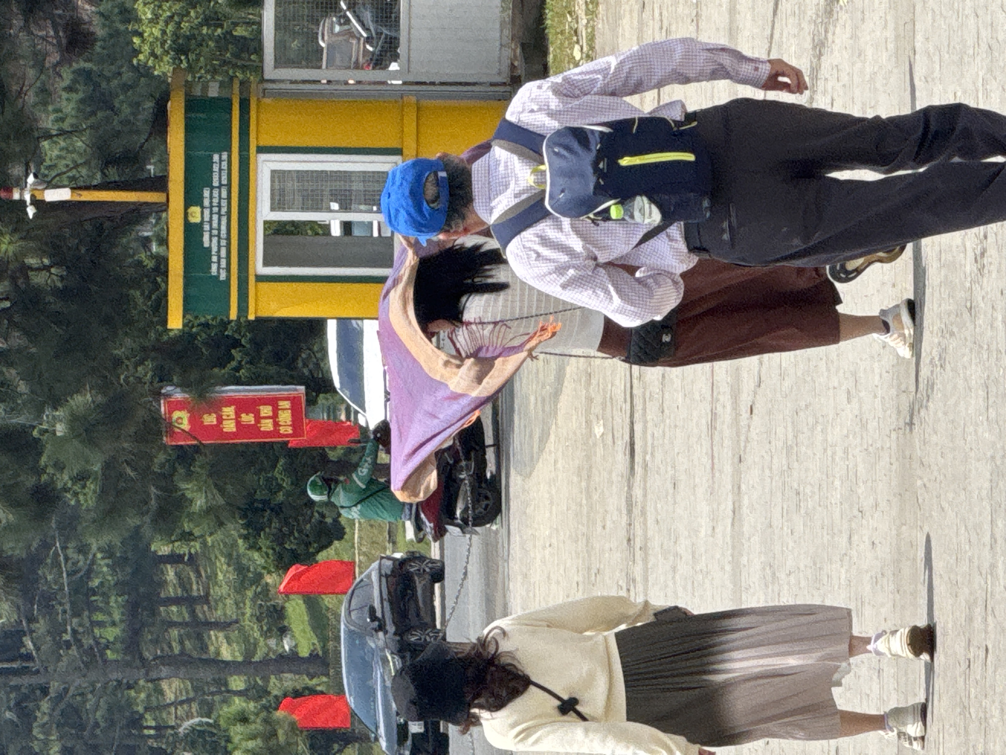 The image depicts a street scene in Da Lat, Vietnam, likely near a checkpoint or entrance, as indicated by a yellow and green booth with signage that appears to be official. In the foreground, three individuals are walking. The person on the left is wearing a black hat, a cream-colored sweater, and a long, pleated skirt that transitions from dark gray to light pink. They are carrying a black crossbody bag. In the center, a person with dark hair is partially obscured by a large purple and beige scarf being held up. They are wearing a striped top and a brown skirt. To the right, a man wearing a blue baseball cap, a pink and white checkered shirt, and dark pants is walking away from the camera. He has a dark blue backpack with yellow accents, and a water bottle is attached to the side. In the middle ground, a person in a green uniform and helmet is riding a red scooter, and they appear to be holding onto or near the scarf that is covering the person in the center. Several red flags are visible, some mounted on poles, and others potentially attached to vehicles or structures. A black car is parked on the left, and other vehicles and a chain-link fence are visible in the background. The environment suggests an outdoor, possibly slightly hilly area, with trees and vegetation visible behind the structures. The ground is paved with concrete or stone. The overall atmosphere is that of a public thoroughfare on a bright, sunny day.