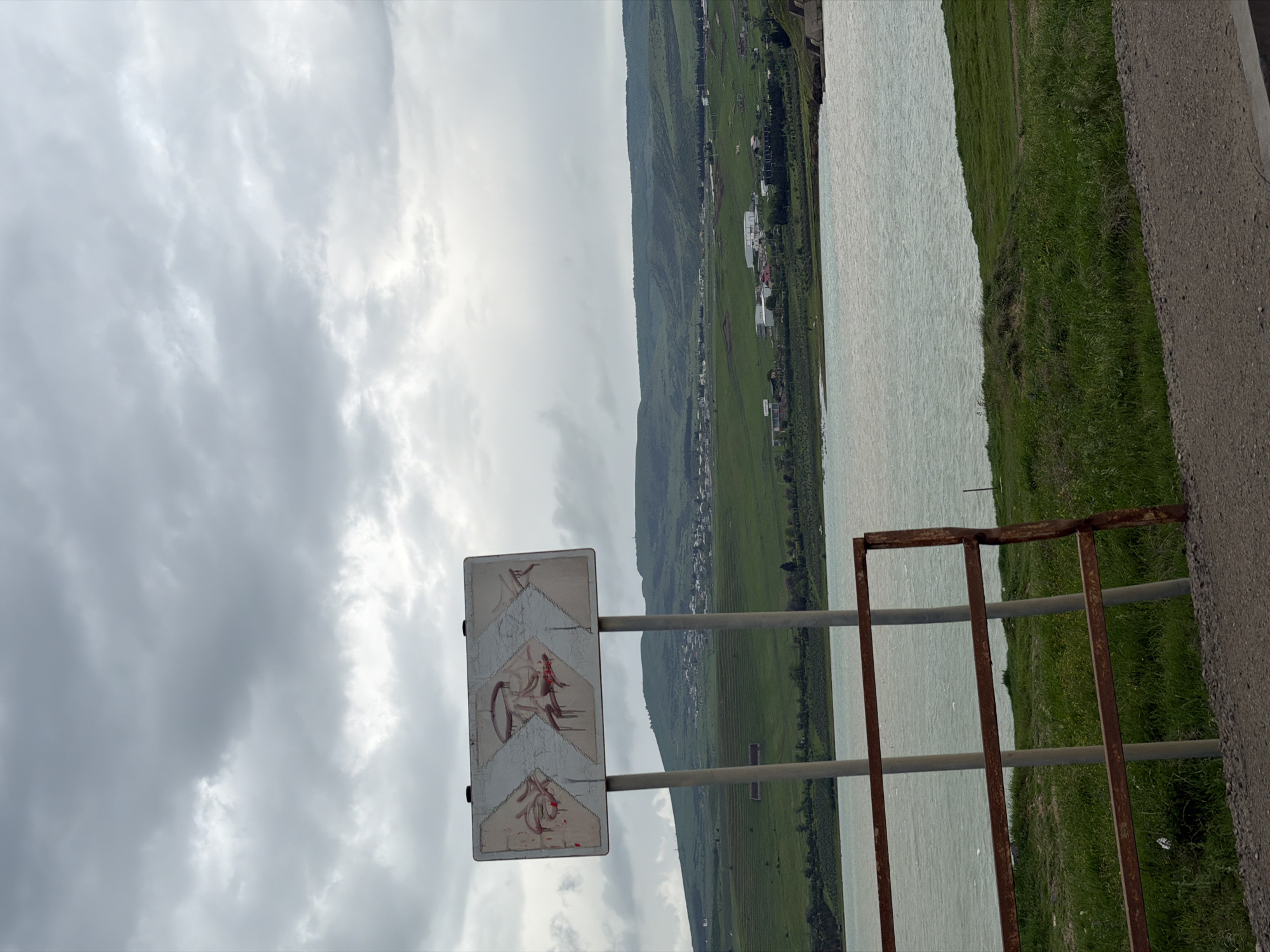 An outdoor shot depicts a landscape with a partly cloudy sky. A road runs horizontally across the lower right, with green grass between the road and a body of water. A rusted metal railing separates the road from the water. On the left side of the road, a road sign stands. The sign is weathered, with what appears to be an image of bicycles. In the background is a green, hilly landscape dotted with structures. The location is likely in the outskirts of Tbilisi, Georgia, where similar landscapes can be found.