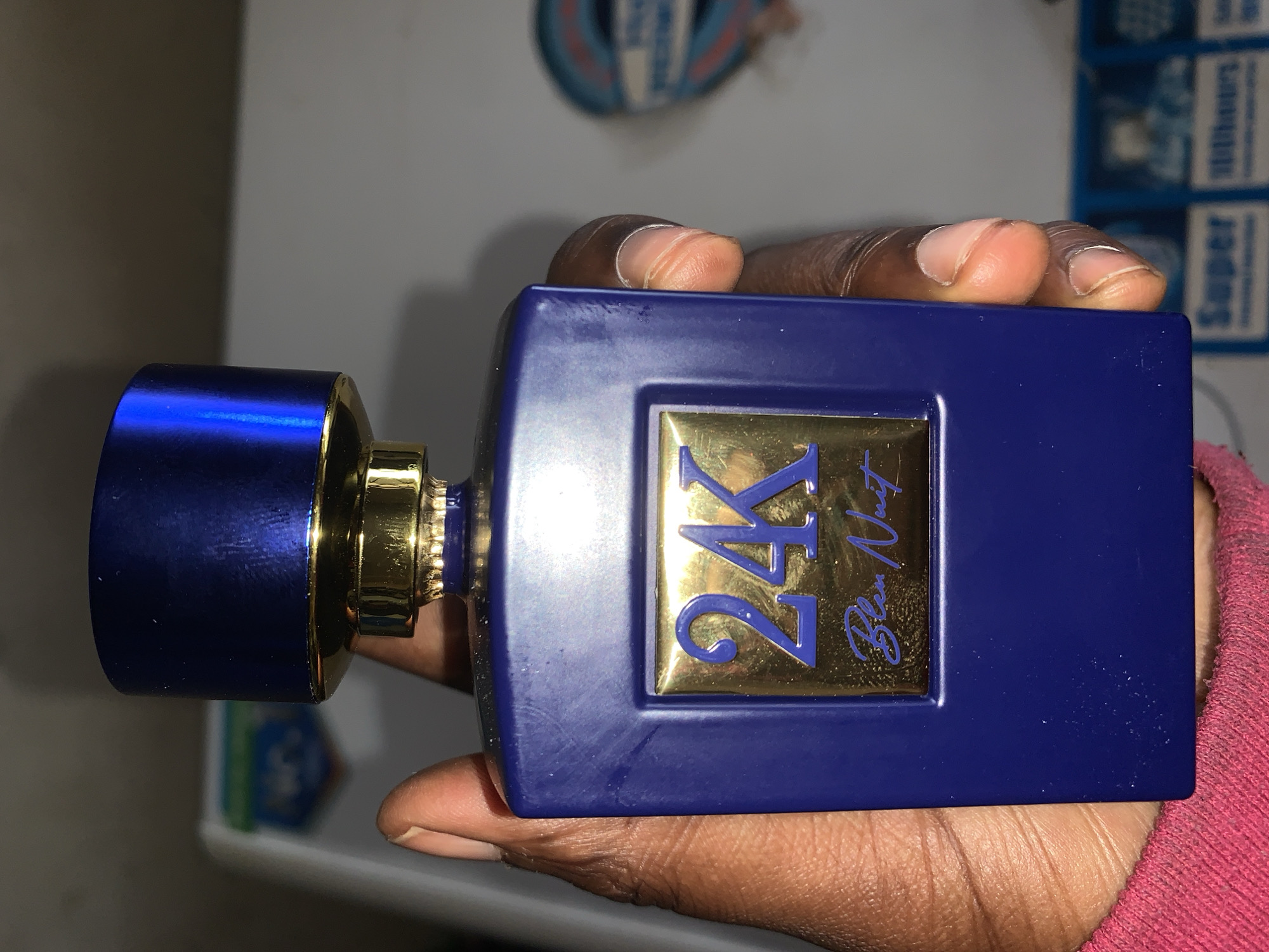 An overhead shot captures a dark-skinned hand holding a rectangular, deep blue perfume bottle with gold accents. The hand, with visible fingernails, grasps the bottle firmly. The perfume bottle features "24K" prominently in large gold characters with a blue outline on its front, beneath which "Bleu Nuit" is written in elegant blue script. The cap of the bottle is a matching metallic blue, connected by a ribbed gold collar.
The scene appears to be indoors, possibly on a table or counter, with the background blurred. In the soft-focused background, hints of other objects are visible, including what looks like a blue and white patterned item (possibly a shoe) and some packaging that reads "Super" and "Lithium," suggesting battery packs. The lighting is diffused, indicating an indoor setting. No specific activity beyond holding the bottle is discernible, perhaps for display or inspection. The photograph is situated in the context of New Stadium, Nigeria.