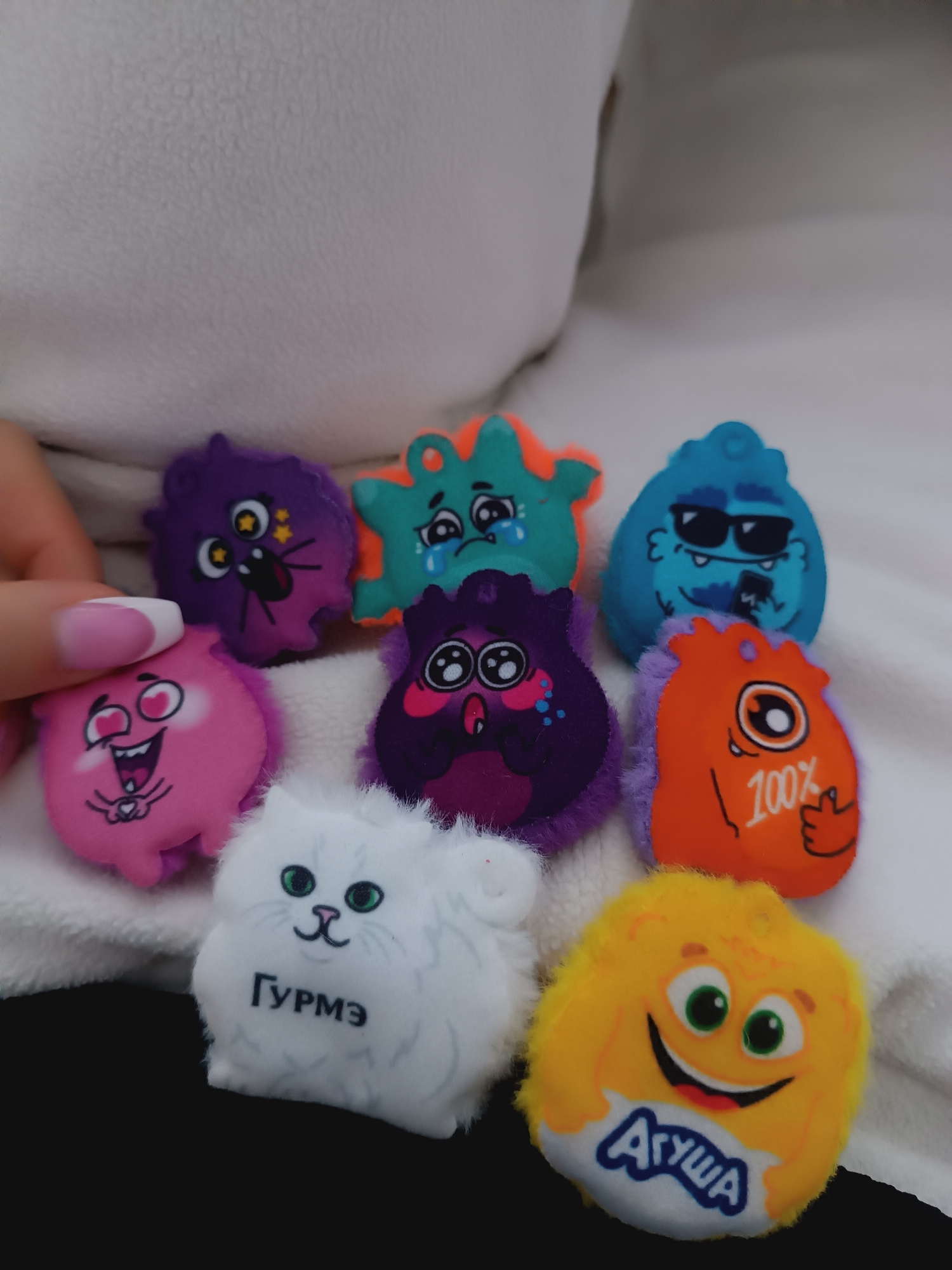 The image shows a close-up of eight small, cartoon-like plush toys arranged on a white, textured surface. A hand with manicured nails is partially visible on the left side of the image. The toys vary in color and design, including purple, green, blue, pink, orange, white, and yellow figures, each with different facial expressions and some with printed text. The text on the white toy reads "ГУРМЭ", while the yellow toy has the word "АГУША" printed on it. The toys are set against a blurred background, possibly a white pillow or blanket, on top of a black surface. The location is Unknown, Unknown.