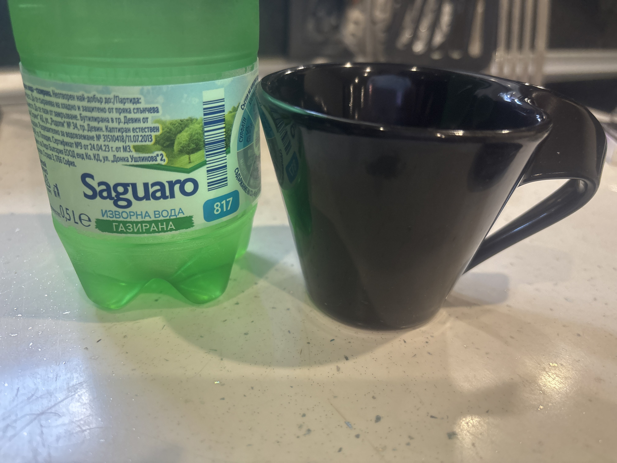 The image shows a close-up, slightly overhead view of a green plastic bottle of Saguaro mineral water and a black ceramic mug on a speckled countertop. The bottle is on the left side of the frame, and the mug is on the right. The bottle's label is partially visible, displaying text in Bulgarian, including "Saguaro" and "ИЗВОРНА ВОДА ГАЗИРАНА" (Spring Water Carbonated). A barcode is also visible on the bottle. The black mug has a curved handle and appears to be empty. The countertop is light-colored with dark speckles and some scattered small debris. The lighting suggests an indoor environment. There are no people visible in the image. The provided context mentions Burgas, Bulgaria, but the image itself does not contain any visual cues to confirm this specific location.