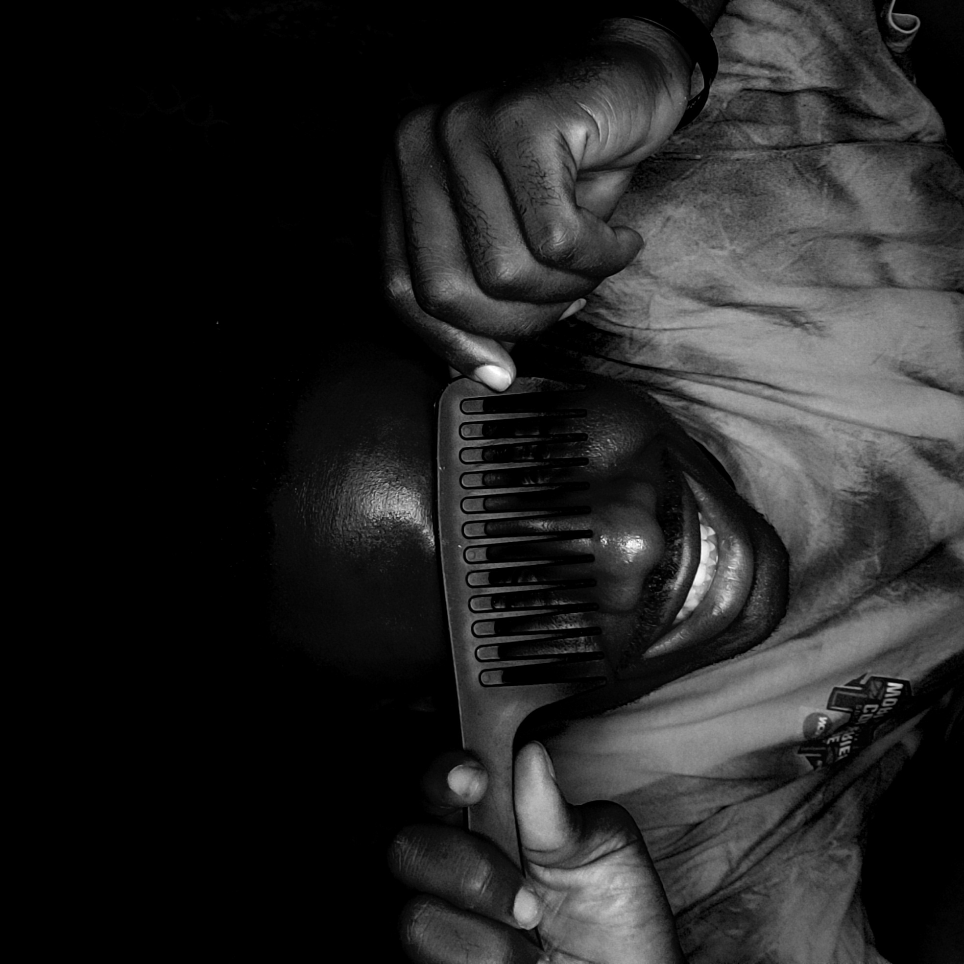 A black and white, close-up portrait shows a person partially obscuring their face with a large, wide-toothed comb. The image is rotated, with the person's head positioned diagonally. The subject, who has dark skin, is smiling broadly, revealing their teeth, conveying a sense of playfulness or joy. Only the lower part of their face, including the mouth and chin, is clearly visible behind the comb's teeth, along with portions of their forehead and cheek. Their eyes are obscured by the comb.
One hand holds the comb near the bottom right, with fingers gripping the handle, while another hand is visible near the top left, likely part of the same person. A dark wristband is visible on the person's upper arm or wrist on the left side. The lighting is dramatic, highlighting the contours of the skin and the texture of the comb, while casting deep shadows that create strong contrast with the illuminated features.
The subject is wearing clothing or covered by fabric, which is visible on the right side of the image, showing wrinkles and folds. On this fabric, near the bottom right, an upside-down label or design is visible with partially decipherable text. Parts of the text include "MOW" at the top, followed by "CO" and "WIE" or "WWE" below it, in a stylized, blocky font. The overall setting appears to be an interior in Narayi, Nigeria, with a dark, indistinct background.