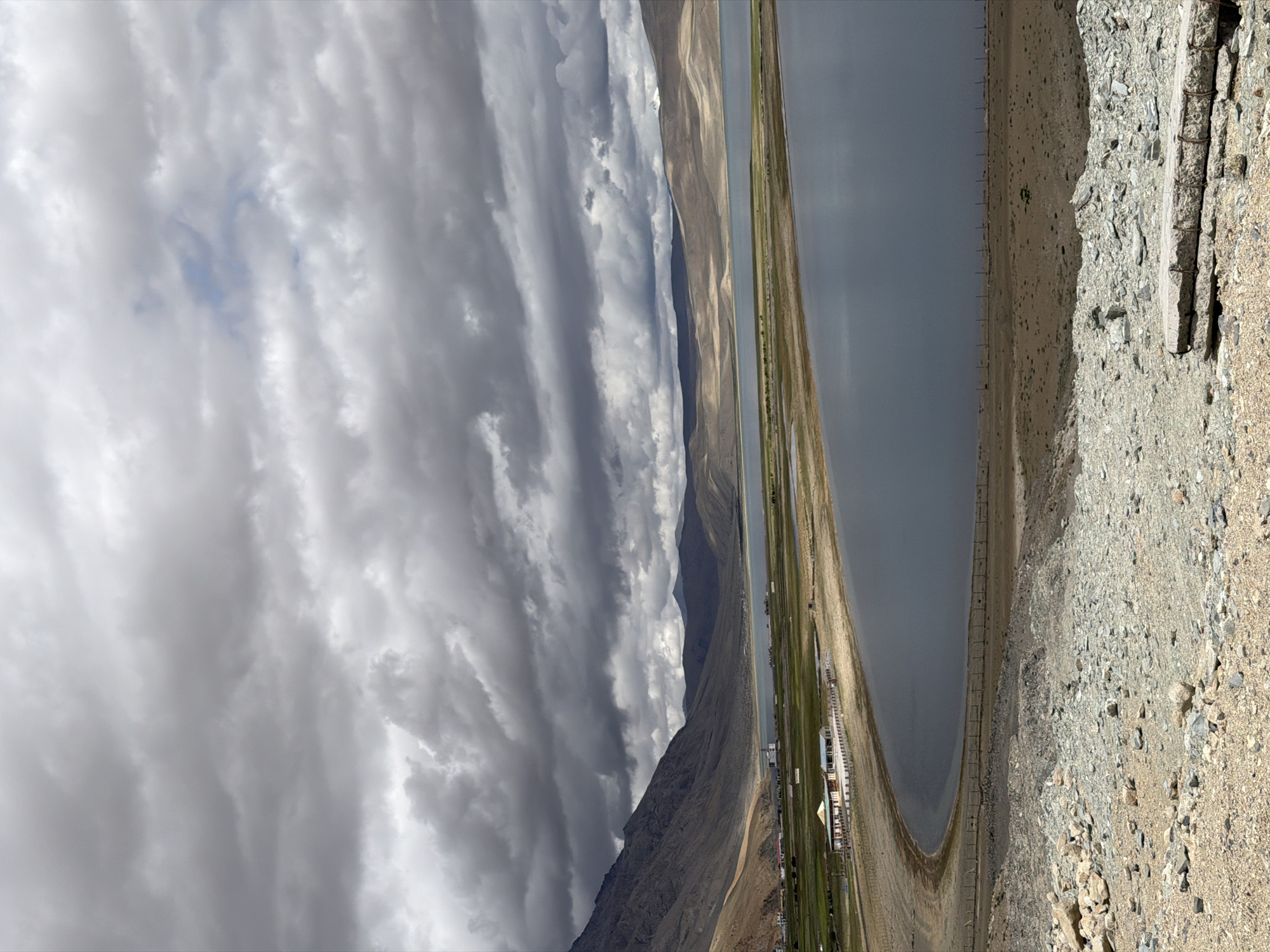 The image shows a wide panoramic view of the Tso Kar lake in Karzok, India, under a cloudy sky. In the foreground, there is a rocky, desolate landscape. A path or road made of gravel and scattered rocks leads down towards the lake. Along the edge of the lake, there is a sandy shore lined with a simple fence. In the middle ground, a long, rectangular building with a light-colored roof and several smaller attached structures is situated on the shore, surrounded by green fields. Further in the distance, the lake stretches out, meeting a plain dotted with some scattered structures and then the vast, barren mountains. The sky is filled with dramatic, dark grey clouds, suggesting an overcast or impending rainy weather. The overall impression is one of a remote, high-altitude landscape with a hint of human settlement near the water.