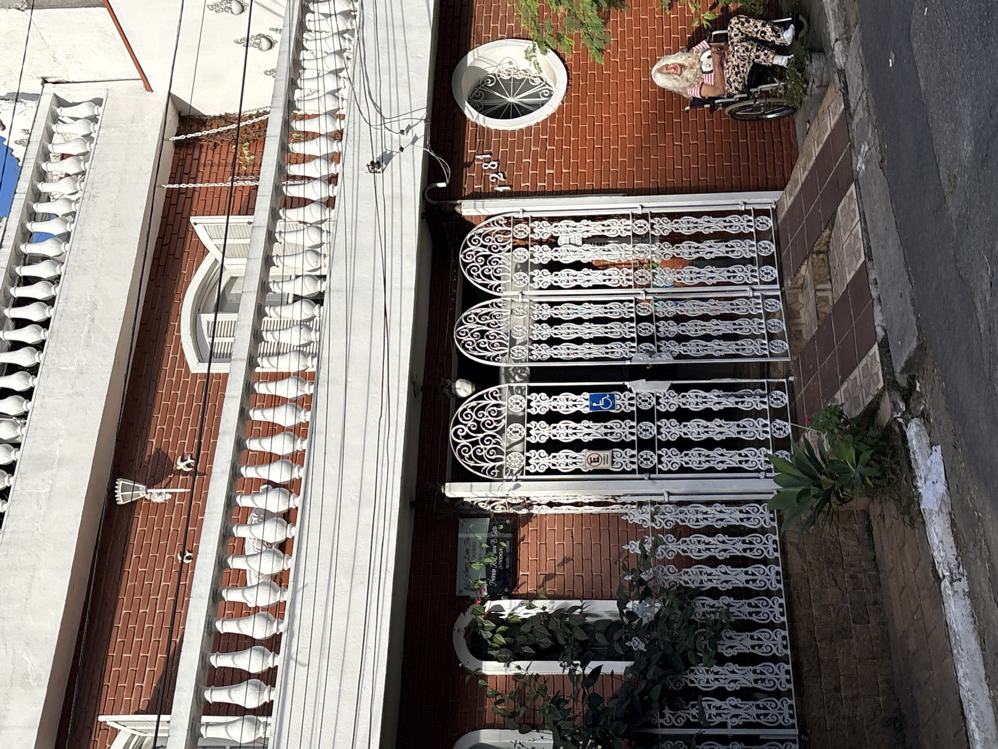 The image shows a brick building with white railings and decorative gates. A woman with white hair, wearing a striped shirt and patterned pants, is seated in a wheelchair to the right of the entrance. She is looking at the camera. There is a ramp leading up to the ornate white gates. A sign with text and symbols is visible on the left side of the entrance. The building is situated on a street in what appears to be a residential or commercial area. The architecture suggests a location in São Paulo, Brazil.