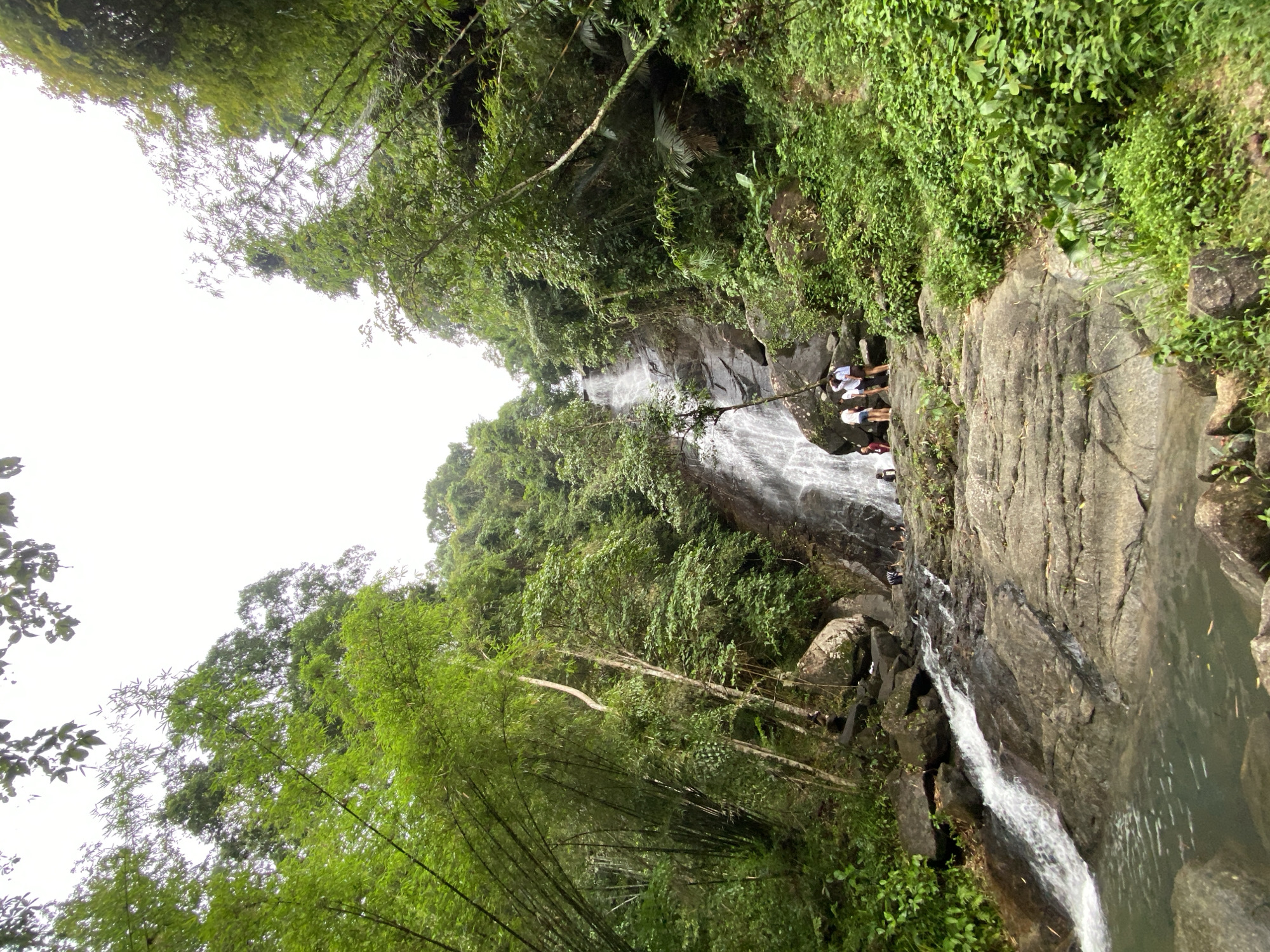 The image shows a waterfall in Ban Bang Khli, Thailand. The waterfall is surrounded by lush green vegetation. Several people stand on the rocks near the waterfall's edge. Water cascades down the rocks, forming pools at the base. The sky is overcast.
