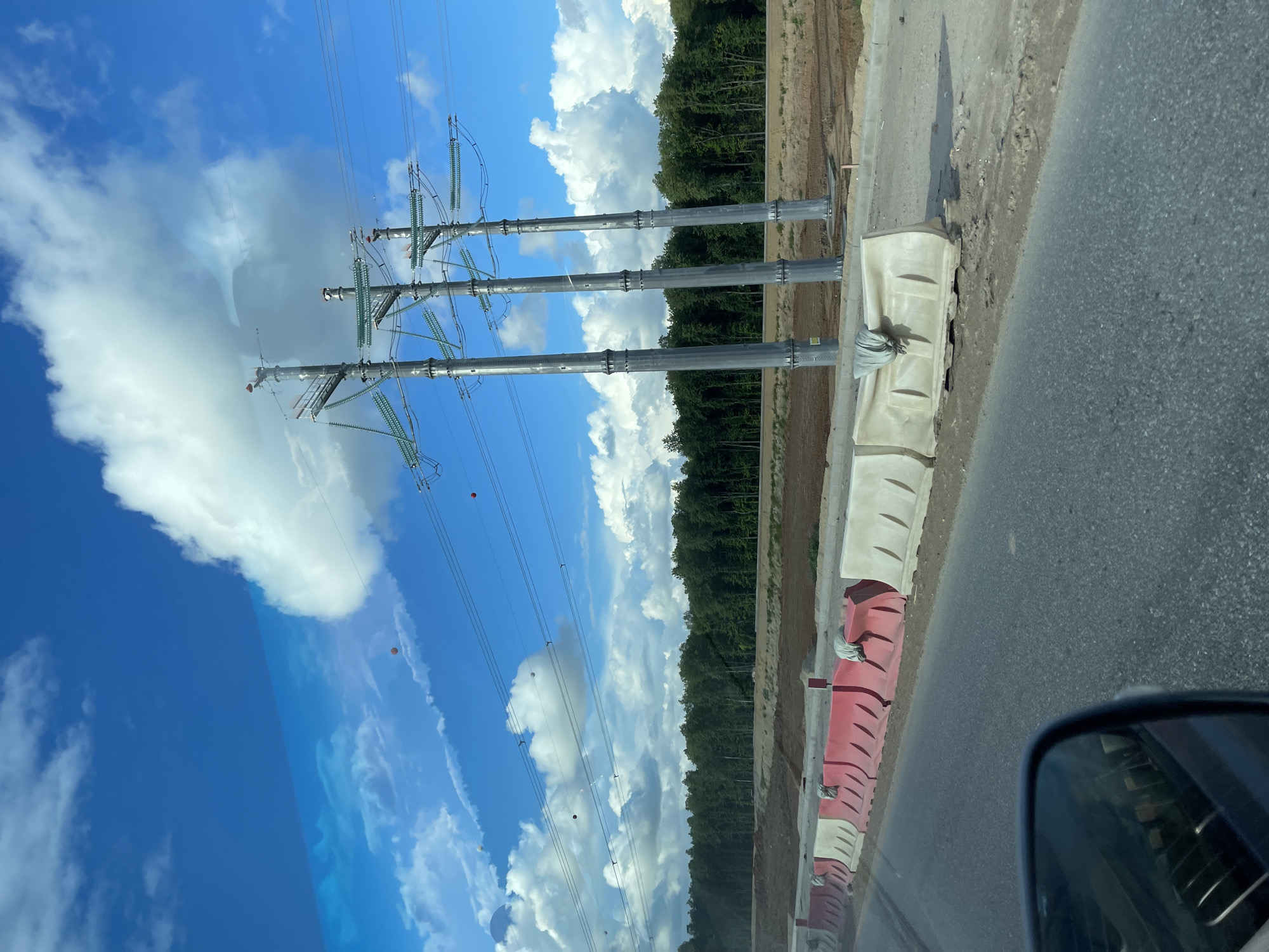 The image shows a row of three high-voltage power transmission towers erected next to a road in Novodrozhzhino, Russia. A blue sky with cumulus clouds forms the backdrop. The towers appear to be newly installed, and power lines are strung between them. Concrete barriers painted red and white line the edge of the road. The road is asphalt, and a forest borders the opposite side of the towers. The view is taken from inside a vehicle, as evidenced by the side-view mirror visible in the lower right corner of the image.