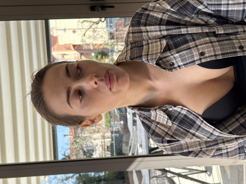 A medium-shot portrait captures an adult female indoors, looking out through a large window in Albisano, Italy. The female, positioned off-center, has light skin and her hair pulled back. She is wearing an open long-sleeved plaid shirt, featuring a pattern of blue and beige squares, which reveals a black bra underneath. Her head is slightly tilted, and her gaze is directed towards the lower right of the frame, past the viewer. Strong sunlight illuminates the left side of her face and shoulder. The background, visible through the window and slightly out of focus, shows an outdoor environment. This includes several residential buildings with terracotta-colored roofs and light-toned walls, characteristic of Mediterranean architecture. In the mid-ground, an outdoor patio or terrace area is visible, furnished with dark-colored tables and chairs. Sparse vegetation is present, and a clear blue sky is visible at the top. Horizontal window blinds are partially visible at the uppermost edge of the window frame.