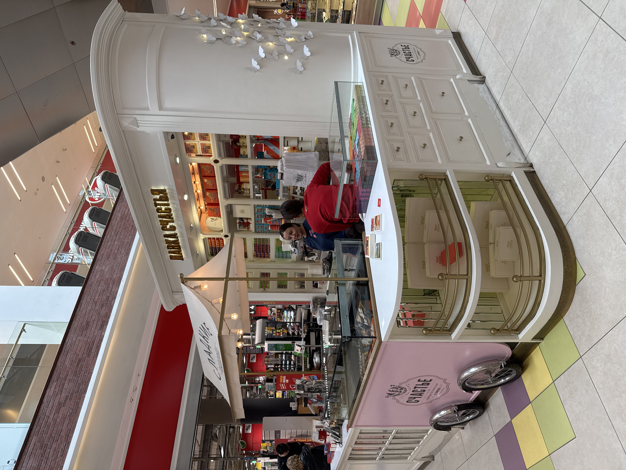 A brightly lit retail kiosk, designed in white with decorative gold accents, stands in an indoor shopping environment. The kiosk, likely a confectionery or chocolate shop, features a curved roofline and elaborate moldings. White paper butterflies are decoratively affixed to its upper facade.
Behind a clear glass display case filled with colorful chocolates or sweets, two employees are visible. One employee wears a red top and stands centrally, while another in a blue top is partially seen. The activity appears to be standard retail operations, with employees ready to serve. A third person, possibly a customer, is partially visible in the bottom left corner.
The surrounding scene includes a multi-colored tiled floor with patterns of white, light grey, yellow, and green squares. In the background, escalators or stairs ascend, flanked by display racks of shoes, further indicating a shopping mall setting. Bright artificial lighting illuminates the entire area.
Notable decorative elements include small, decorative bicycle wheels attached to the lower pink panel of the kiosk. Visible text, primarily in Cyrillic, provides clues about the business. "ЛАВКА СЧАСТЬЯ" (Lavka Schastya), meaning "Shop of Happiness," is prominently displayed on the main signboard. The word "СЧАСТЬЕ" (Schastye), or "Happiness," appears multiple times on the kiosk and its products. Below one instance of "СЧАСТЬЕ," the French phrase "FABRIQUE DE CHOCOLAT" clarifies that it is a chocolate factory or shop. Additionally, "СЛАДКИ" (Sladki), likely part of "СЛАДКИЕ" meaning "sweets," is seen on a canopy structure. The setting is likely in Saint Petersburg, Russia, given the Cyrillic text.