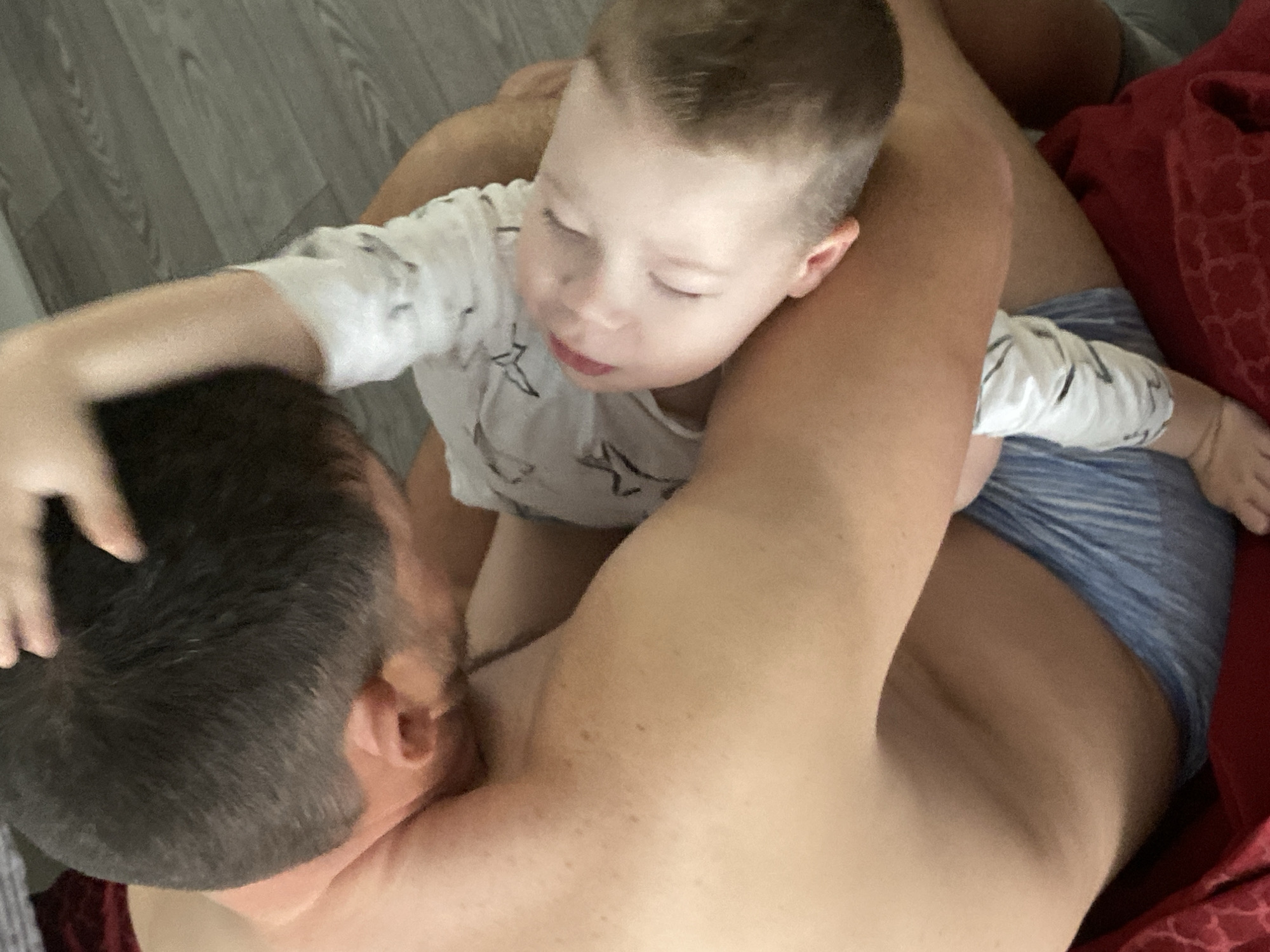 This media file captures an intimate, top-down perspective of an adult and a young child in an indoor setting, identified as Sochi, Russia.
The image prominently features an adult, likely male, lying down with their back and right shoulder in the foreground. They have short, dark hair. A young child, appearing to be a boy, is nestled close to or on top of the adult. The child wears a white long-sleeved top adorned with dark star patterns and blue and white striped shorts or underwear. His eyes are closed or looking downwards, conveying a sense of peacefulness, sleepiness, or deep concentration. One of the child's arms extends around the adult's head or neck area, while the adult's arm is visible embracing the child.
The background shows a light grey wooden floor and a section of red fabric, possibly a blanket or cushion. The scene suggests a tender moment of affection, comfort, or quiet bonding between the two individuals. The lighting indicates an indoor daytime setting. No specific time of day, weather, or visible text can be determined from the image.