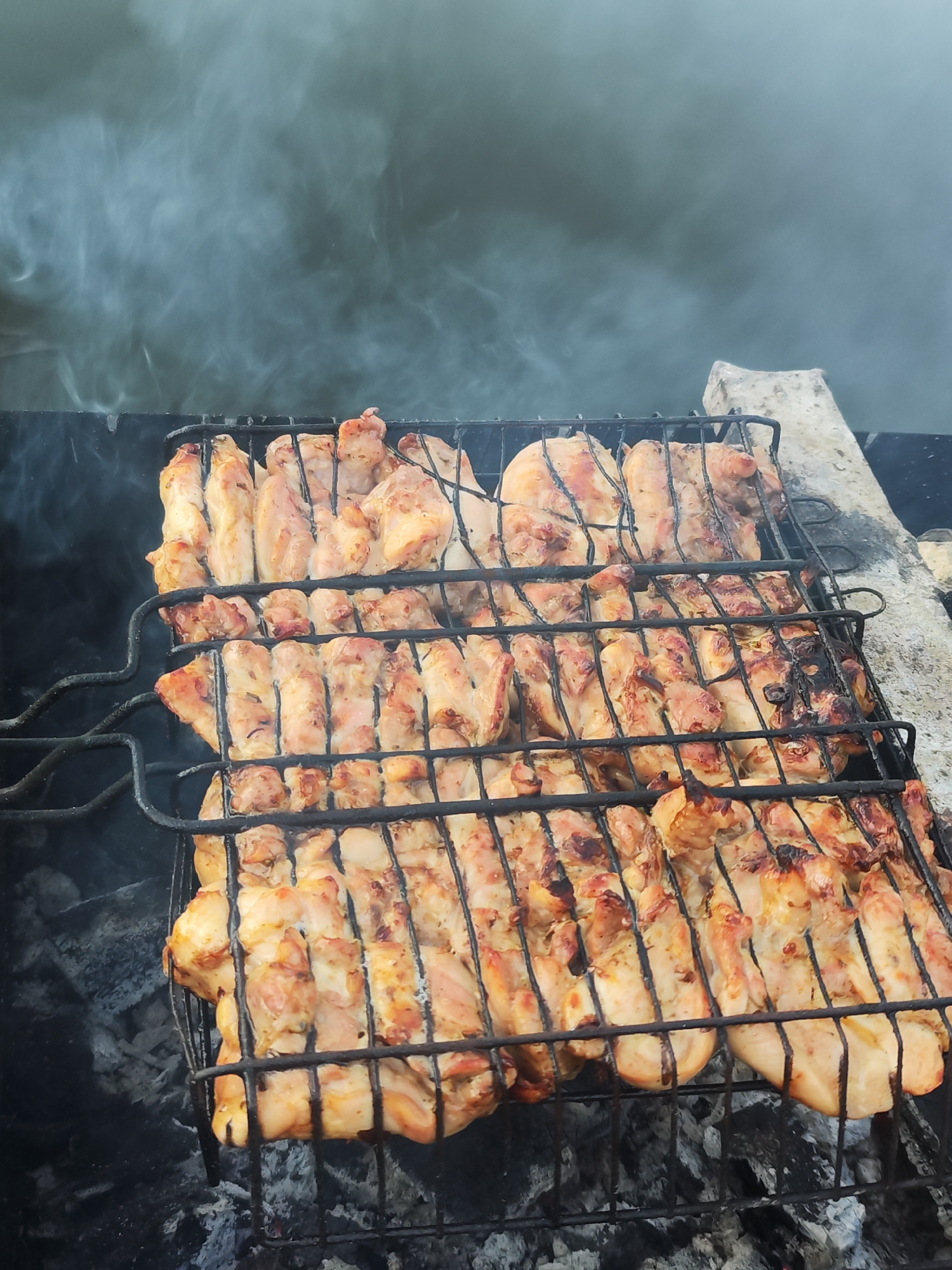 The image depicts chicken pieces being grilled over charcoal in what appears to be an outdoor setting. Smoke is visible in the background. The chicken is contained within a black metal grilling basket. Some pieces exhibit char marks, indicating they have been exposed to direct heat. The environment suggests a possible backyard or picnic area, though specific details about Chaikovsky, Russia, are not discernible from the image.