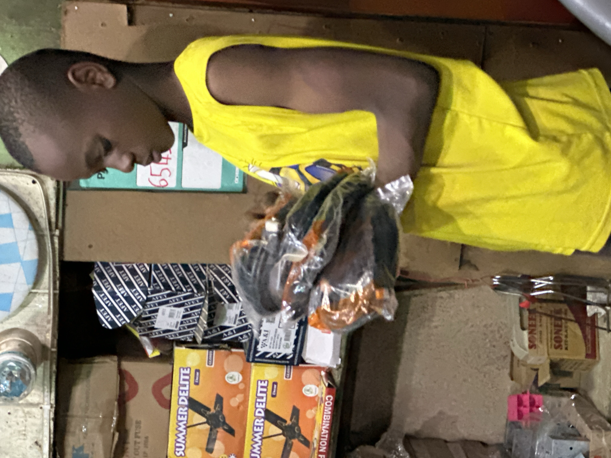 A young boy stands in what appears to be a shop or market stall in Barden Yana, Nigeria. He is wearing a bright yellow sleeveless t-shirt and is holding several items packaged in clear plastic. The items seem to be electronic components or perhaps spare parts, possibly related to appliances given the "SUMMER DELITE" boxes visible in the background, which depict ceiling fans. The background reveals shelves stocked with various goods, including cardboard boxes with labels in English and what looks like the brand "SONEE." The lighting suggests an indoor setting, possibly dimly lit, with some overhead light fixtures visible. The boy's gaze is directed downwards, and his expression is neutral. The overall scene depicts a commercial environment with a focus on the boy and the goods he is handling.