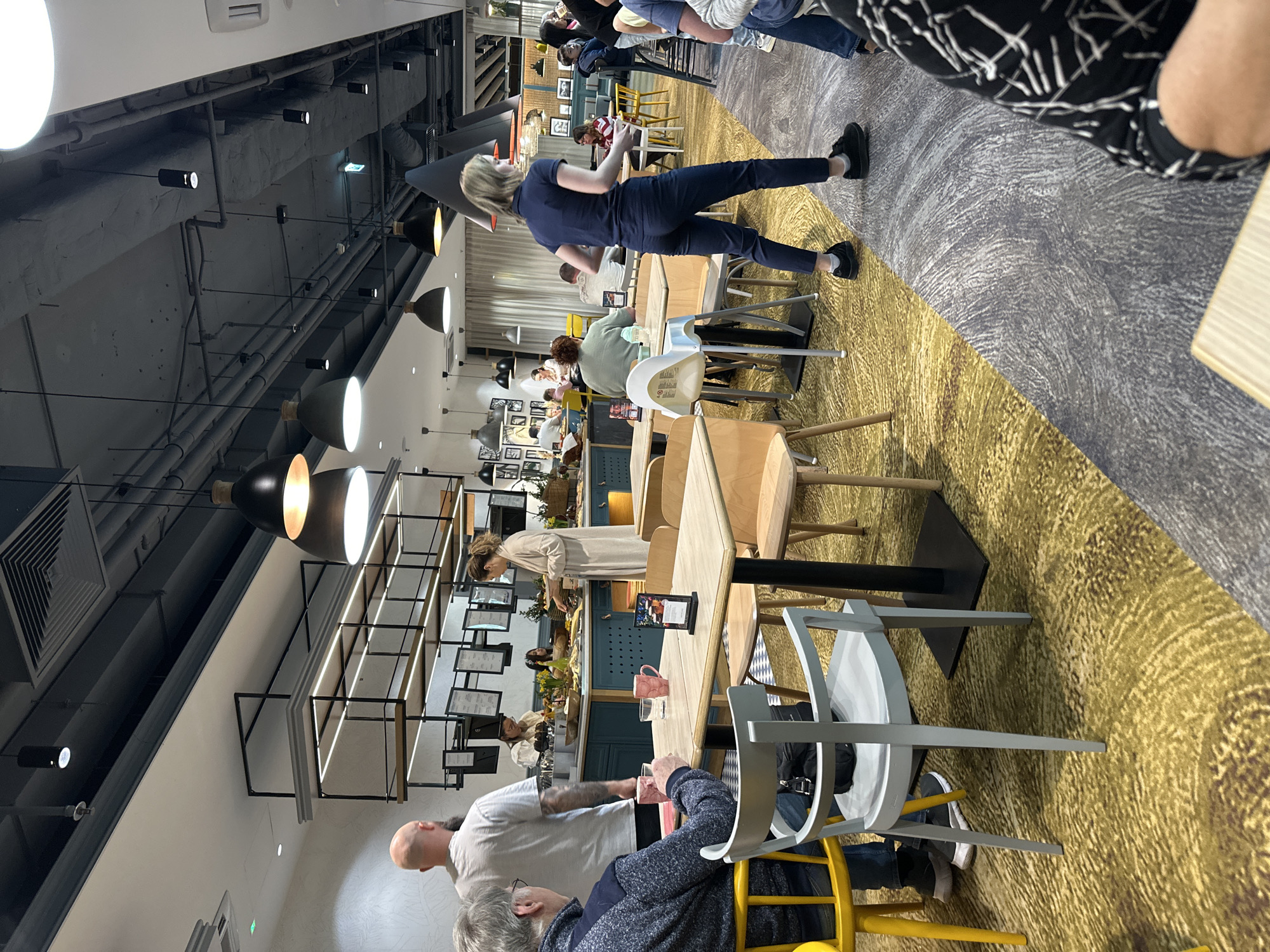 The image depicts the interior of a modern, well-lit cafeteria or restaurant, likely in Wrocław, Poland. Several people are present, some standing and some seated at tables. A woman in a blue shirt and dark pants is walking through the space, possibly toward another table. Seated patrons are interacting with each other. The environment consists of tables and chairs, a patterned carpet, and a buffet or serving area in the background, laden with food. The ceiling features industrial-style lighting fixtures and visible ductwork.