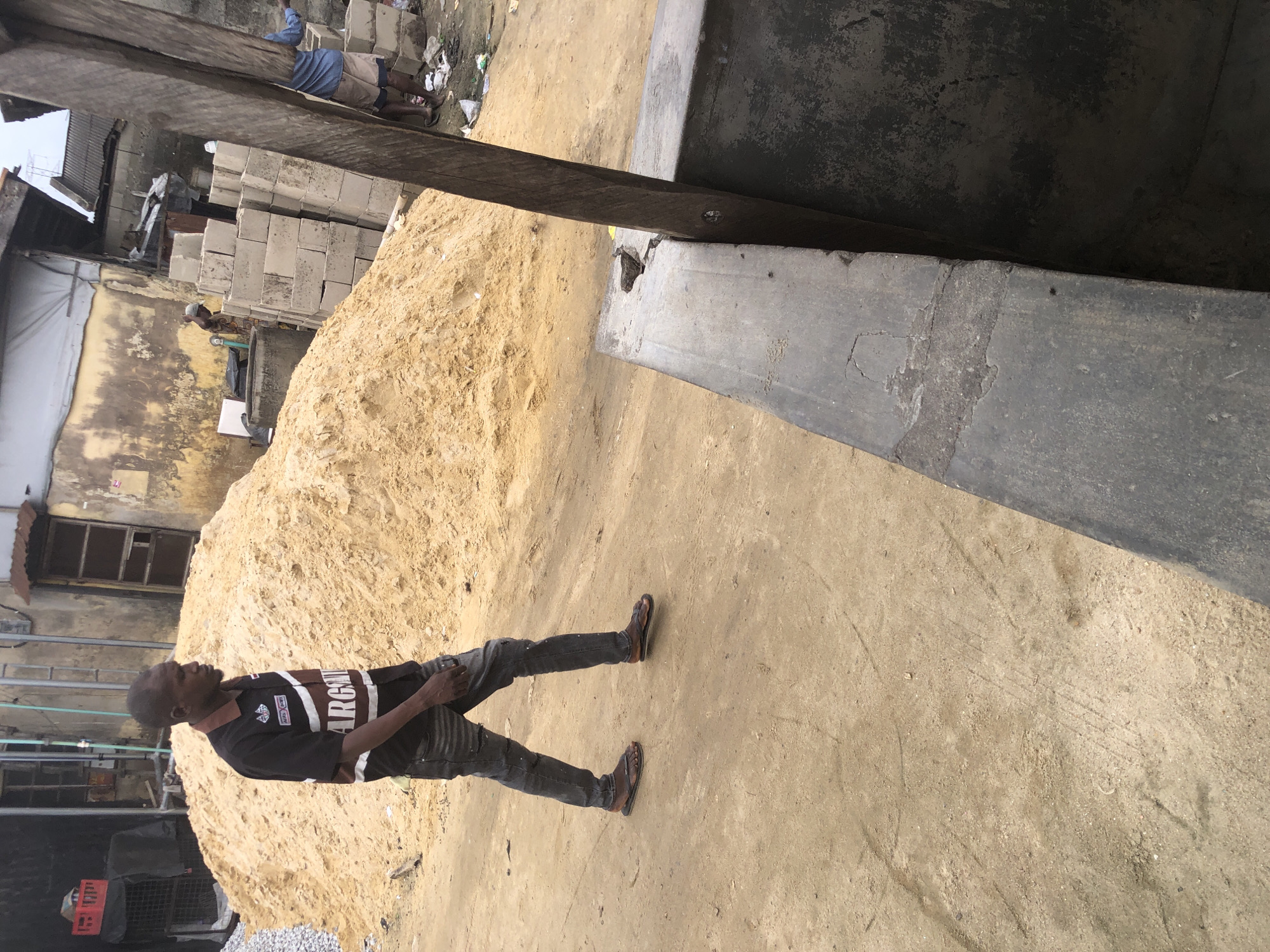 A man walks on a dirt path in Ado, Nigeria. He is wearing a black and maroon collared shirt and dark, ripped jeans. He is also wearing sandals. To his left is a large pile of sand. In the background, there are buildings and a stack of concrete blocks. A wooden beam leans over a concrete structure in the foreground.