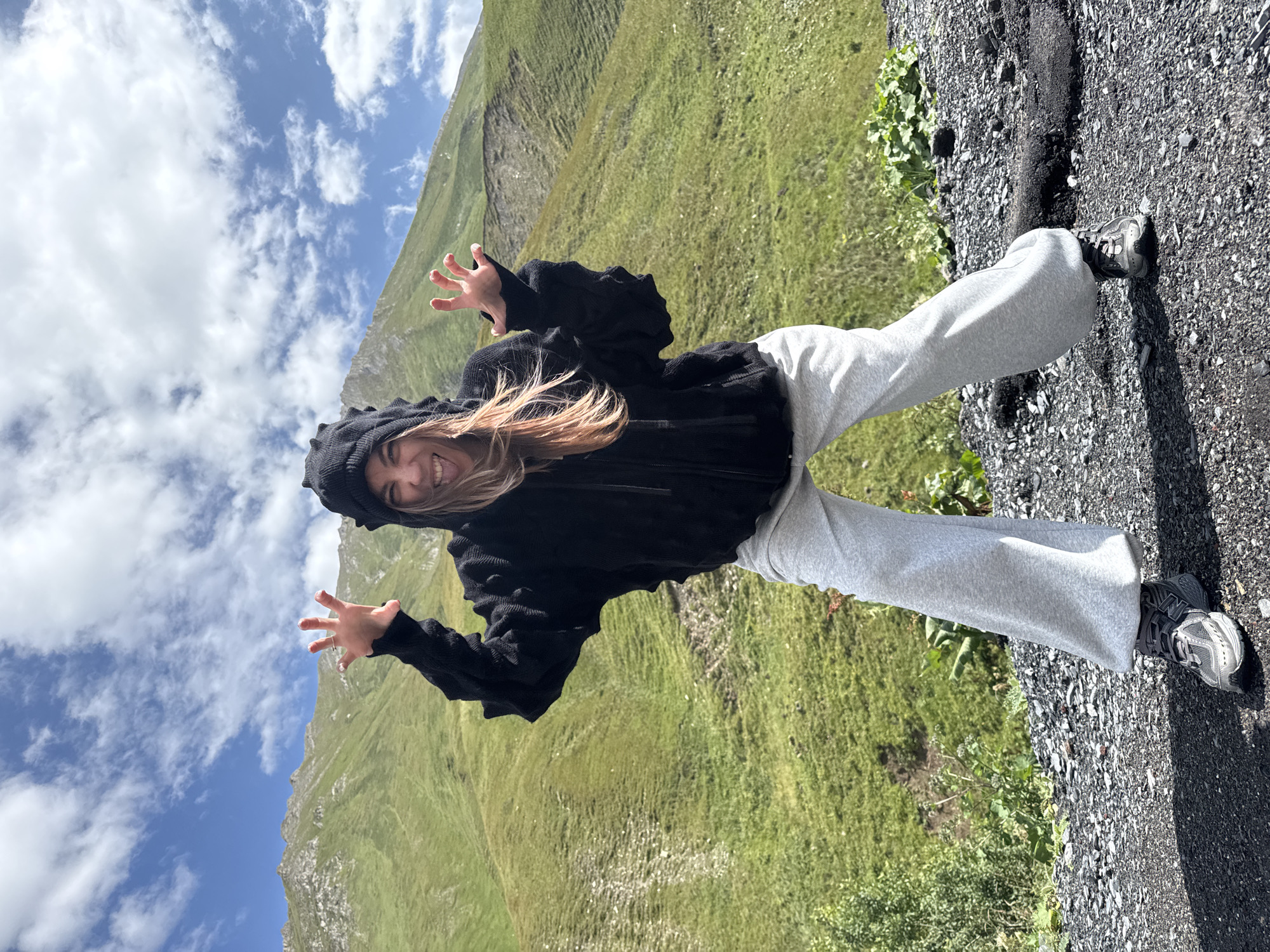 A young woman with light brown hair, wearing a black hooded jacket with dinosaur-like spikes on the hood and light grey sweatpants, stands on a rocky surface in front of a grassy, mountainous landscape. She is making a playful "monster" face with her tongue sticking out and her hands raised with fingers splayed. The background features lush green hills under a blue sky with scattered white clouds. The location is identified as Almasiani, Georgia.