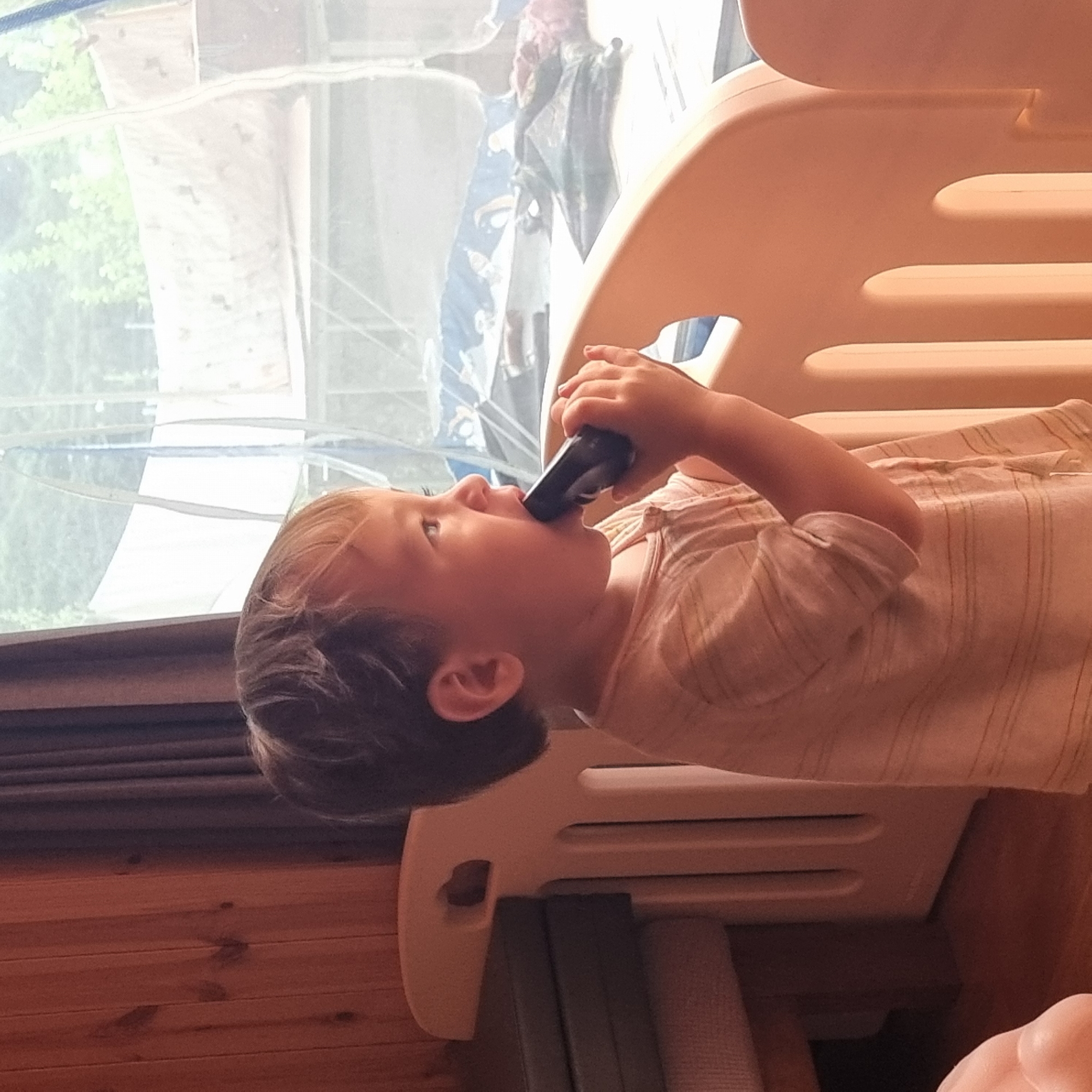 A young child is standing inside a playpen, holding a black object to their mouth. The child is facing to the right and looking out of a window. The playpen is made of light-colored plastic. In the background, there is a window with sheer curtains, revealing trees and possibly a structure outside. The room has wooden paneling on the left side and a dark curtain. The child is wearing a light-colored t-shirt with thin horizontal stripes. The overall setting appears to be indoors, likely a residential area. The image does not contain enough information to determine the specific location in Dunnae-myeon, South Korea.