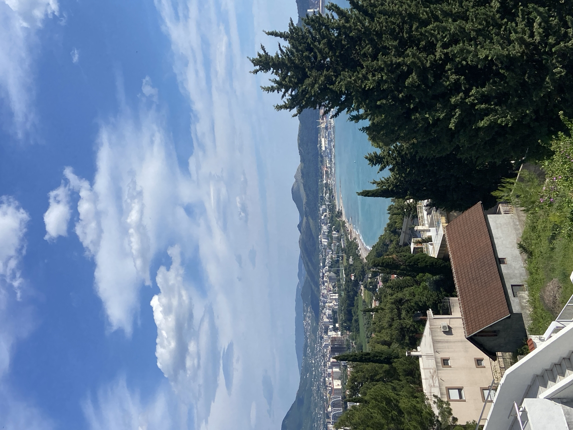 The image shows a high-angle view of Šušanj, Montenegro. In the foreground are the tops of buildings, trees, and a staircase. The middle ground consists of a cityscape stretching along the coastline. Mountains rise in the background and the Adriatic Sea extends to the horizon. The sky is blue with scattered clouds.