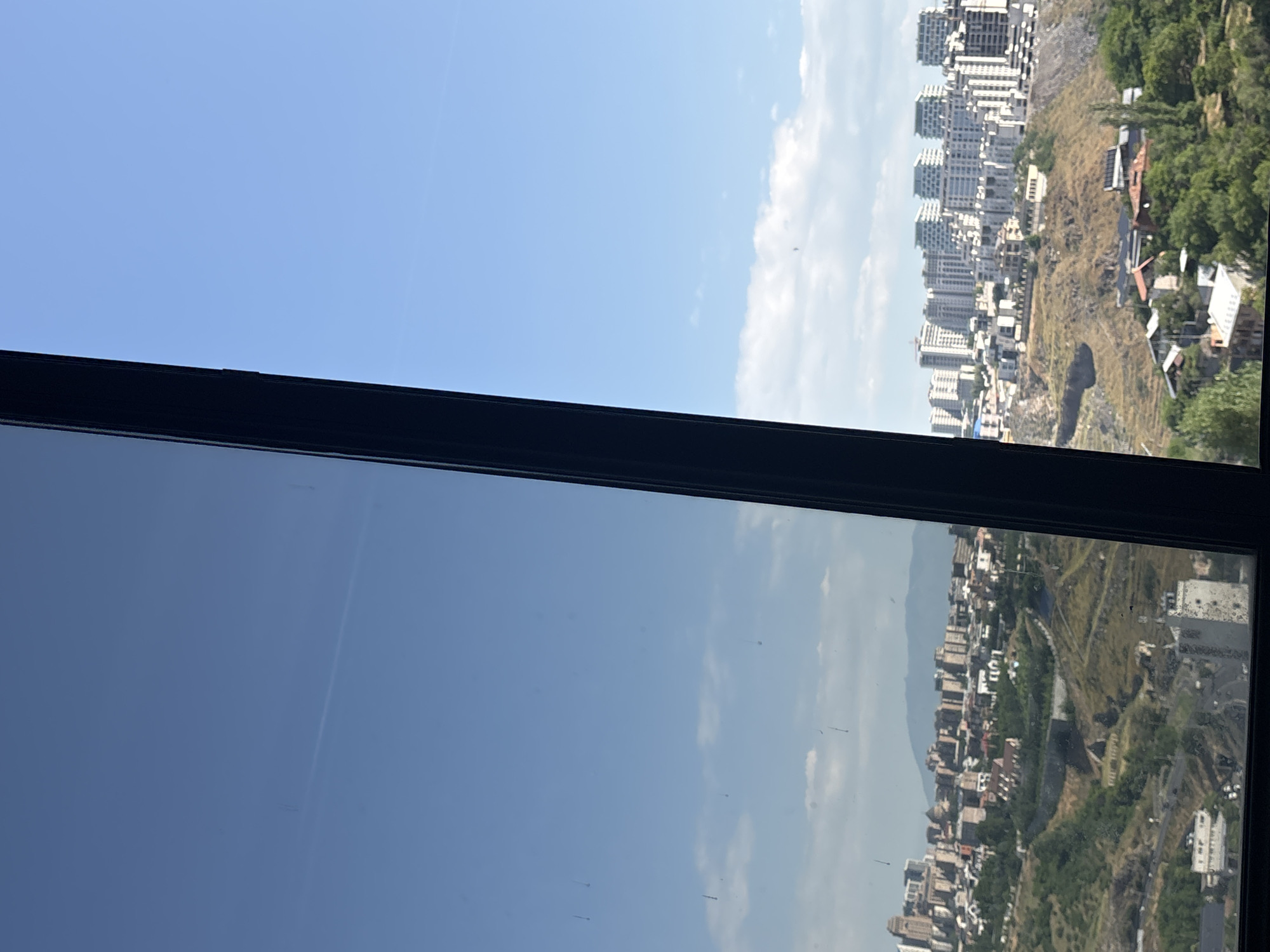 The image provides a panoramic view of Yerevan, Armenia, taken from an elevated position looking across the city. The foreground is dominated by a dark, vertical frame, likely from a window or balcony, which bisects the image.
To the left of the frame, the sky is reflected, showing a clear blue expanse with subtle clouds and a faint contrail from an aircraft. Some smudges or debris are visible on the reflective surface.
To the right of the frame, the actual cityscape of Yerevan unfolds. The scene depicts a dense urban environment characterized by a mix of architectural styles. In the mid-ground, numerous residential buildings, mostly mid-rise and high-rise structures with distinctive white facades and angular roofs, are clustered together on a rolling, hilly terrain. These modern apartment complexes are interspersed with older buildings and visible infrastructure.
The terrain itself is rugged and appears to be partially developed, with slopes covered in scrub vegetation and some trees. Roads and pathways wind through the landscape. Further into the distance, a range of mountains forms a backdrop to the city, partially obscured by atmospheric haze.
The overall impression is of a sunny day with a wide view of a sprawling city situated in a valley surrounded by hills and mountains.