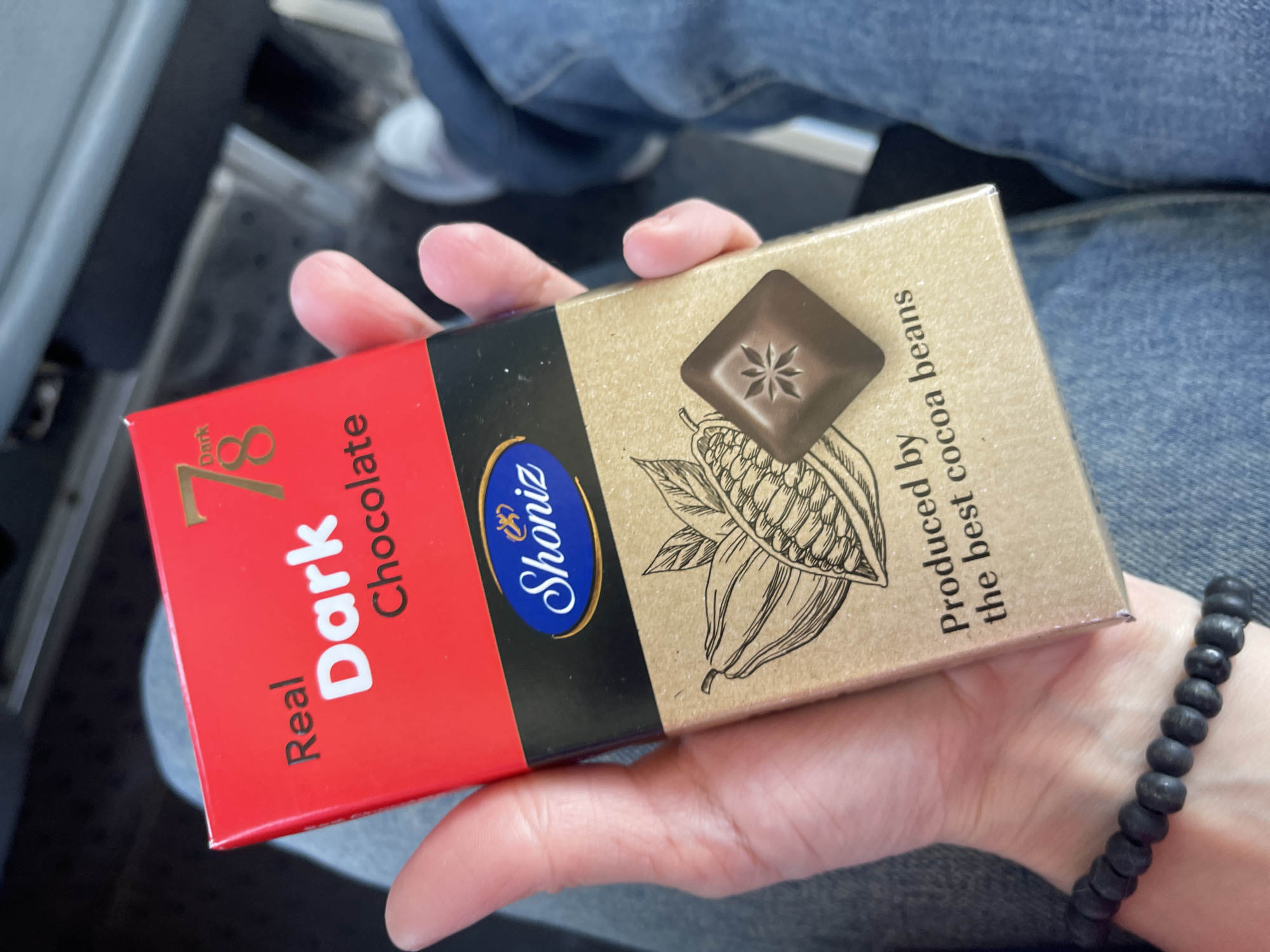 A close-up shows a person's hand holding a rectangular box of "Shoniz" brand dark chocolate. The box has a red, black, and brown color scheme. The red section has "Real Dark Chocolate" and the number "78" printed on it. The black portion contains the brand name, "Shoniz" in a blue oval. The brown section displays a sketch of cocoa beans and a square of chocolate. Text on the brown section states, "Produced by the best cocoa beans." The person is wearing a black beaded bracelet. The background suggests the interior of a vehicle or indoor space, with blue denim clothing and a white sneaker visible. There is no information to suggest the city of Moskovskoe, Russia, as being the location of this image.