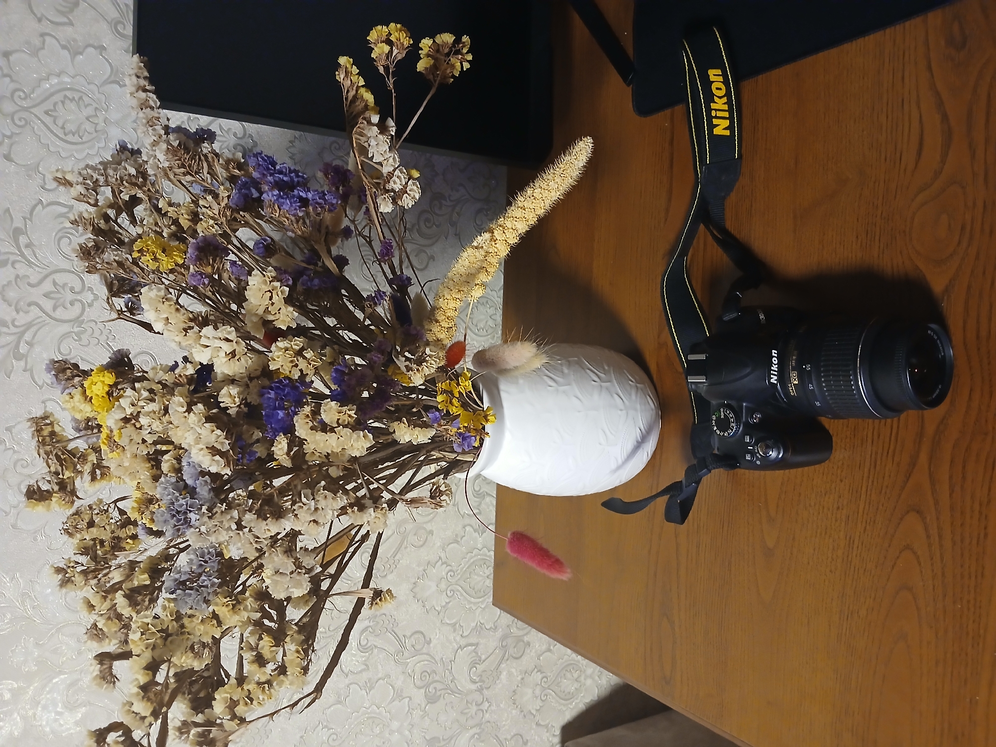 A top-down shot reveals a wooden desk surface bearing a Nikon DSLR camera with its black and yellow strap. Adjacent to the camera, a white vase containing dried flowers in hues of white, yellow, and purple sits. The background consists of a textured wallpaper pattern and a black surface, possibly a monitor or part of the desk's structure. No people are visible. The location is Kamianske, Ukraine.