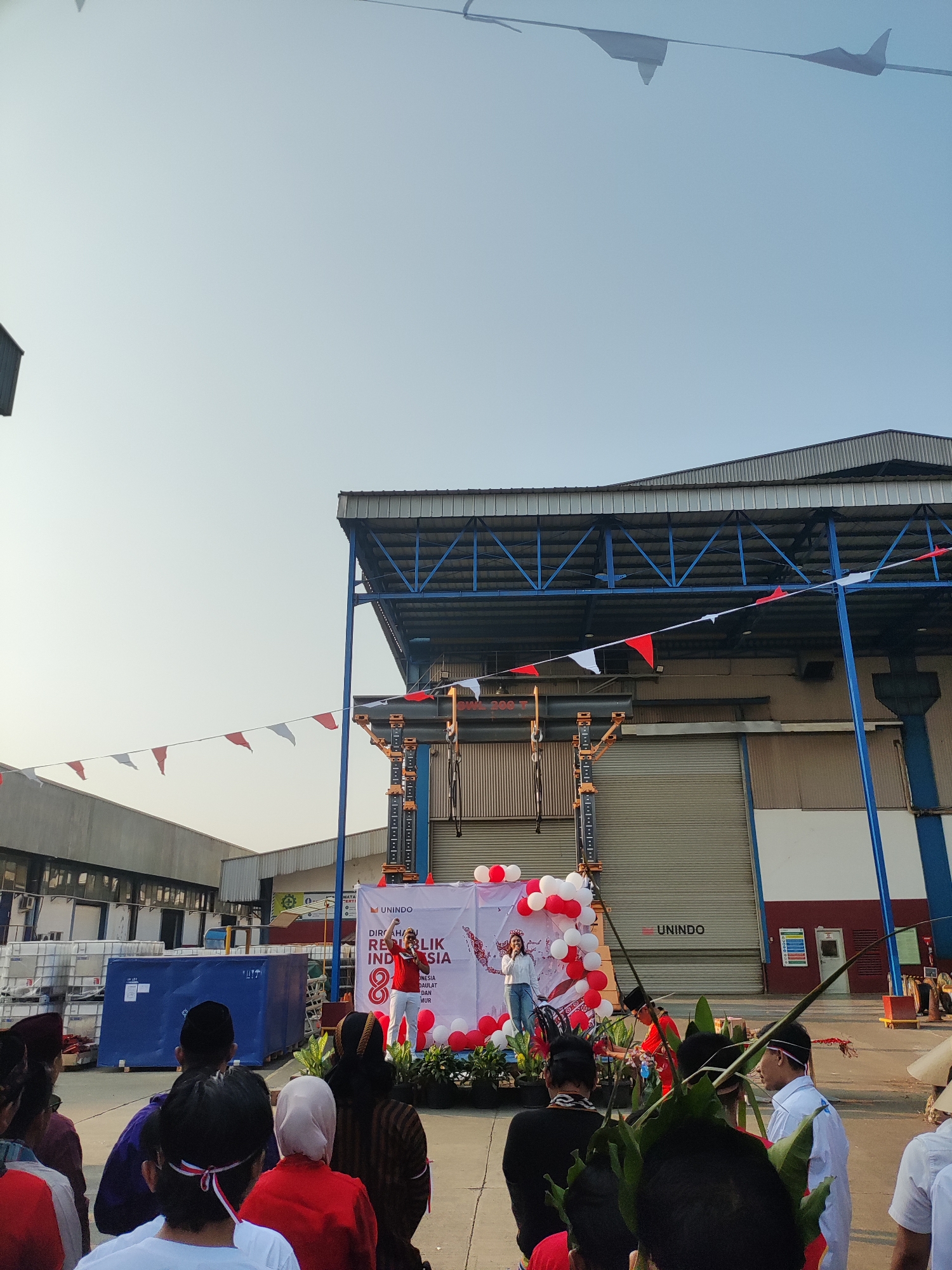 The image depicts an outdoor event in what appears to be an industrial area. In the background, a large warehouse-like building with a corrugated metal roof and light blue structural beams is visible. A large, grey roll-up door is prominent on the building's facade. Red and white triangular flags are strung across the scene, strung between poles and the building.
In the mid-ground, a stage is set up in front of the warehouse. A banner with text and images is displayed on the stage. A young woman in a white top and blue jeans is standing on the stage, holding a microphone and speaking. She is surrounded by red and white balloons. To her right, another individual in a red outfit and hat is performing a dance or ritual. A crowd of people, some wearing traditional Indonesian attire and some in casual clothing, are gathered in the foreground, facing the stage. Some audience members are wearing red and white headbands or sashes.
The ground is paved, suggesting an outdoor industrial or event space. Several large blue containers are stacked to the left of the stage. The sky is clear and pale, indicating daytime. The overall scene suggests a celebration, possibly related to Indonesian Independence Day, given the red and white decorations and the banner. The presence of the UNINDO logo on the banner and building indicates this is likely at a UNINDO facility. While the specific location within East Jakarta is not visually identifiable, the industrial setting and the event suggest it is an industrial complex within the city.