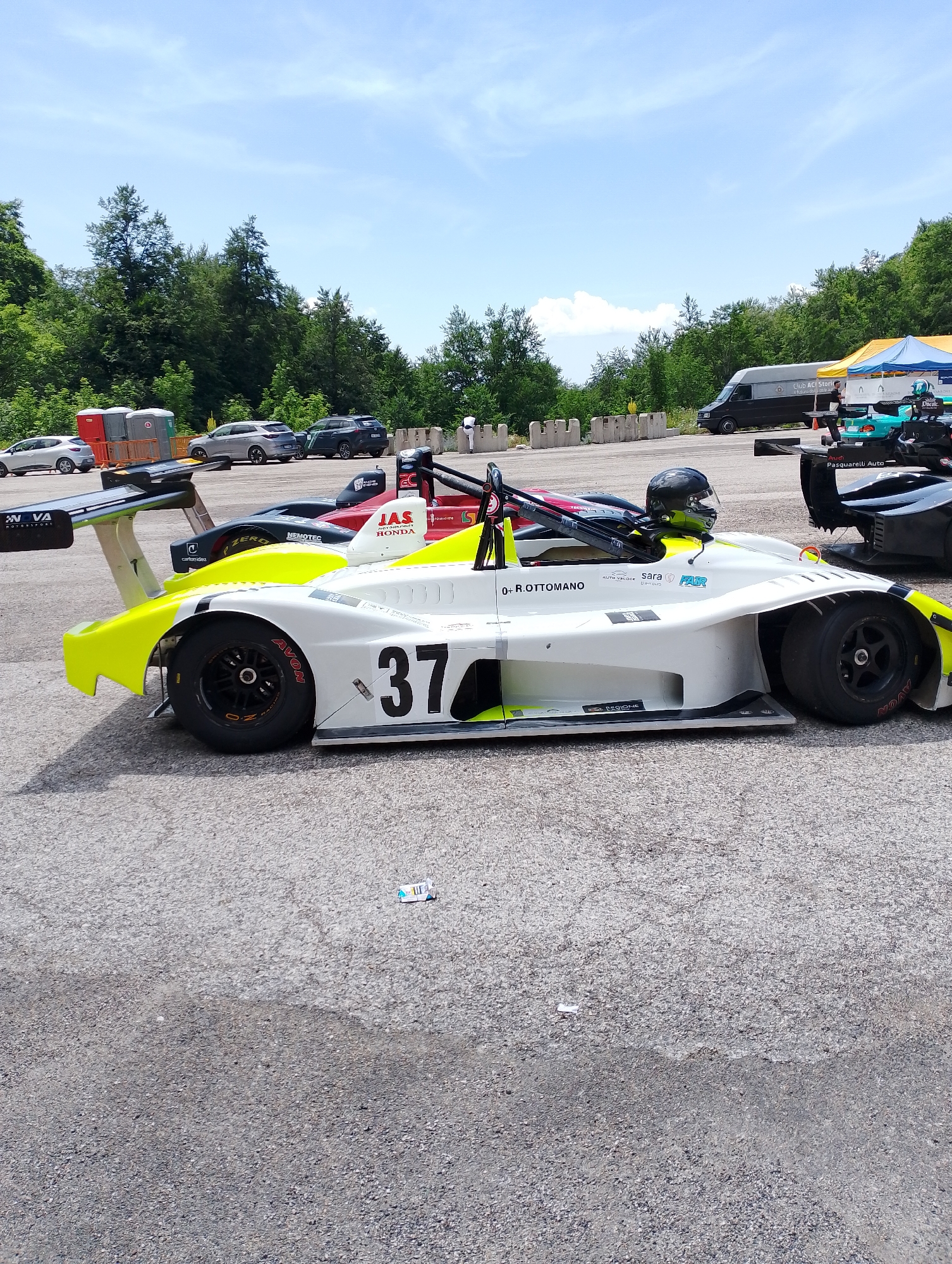 The image shows a race car with the number 37 on its side, parked on an asphalt surface, likely a racetrack. The car is white with yellow accents and has a driver's helmet inside. In the background, there are trees and a cloudy sky. Several other vehicles, including a van and some passenger cars, are visible further back. The scene appears to be outdoors on a sunny day. There is no indication in the image to suggest the location is the city of Rieti, Italy.