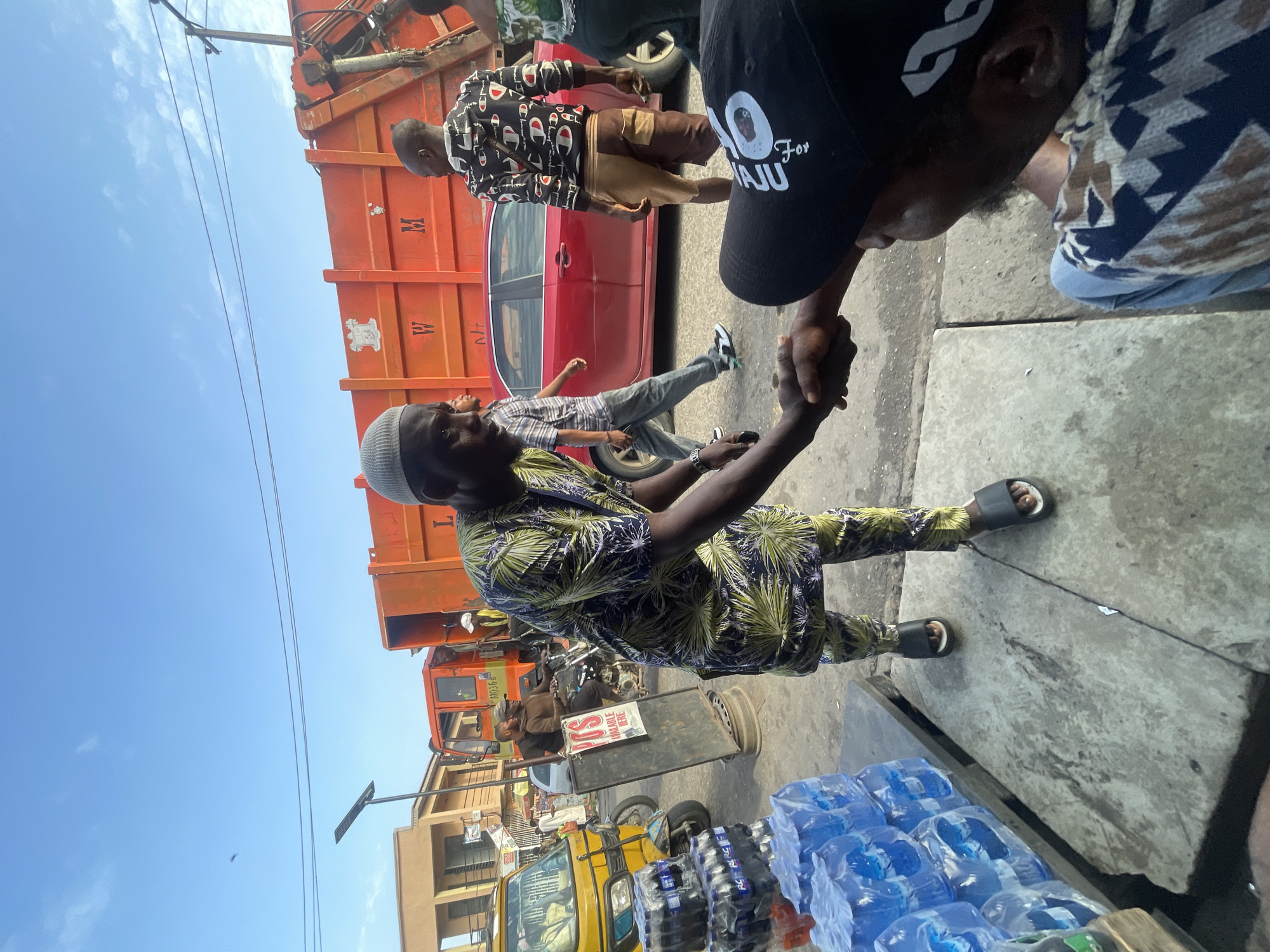 In Akowonjo, Nigeria, a man in a green and blue patterned traditional outfit is shaking hands with another man wearing a black cap and a patterned shirt. The handshake is the central focus, occurring outdoors on a paved surface. Behind them, an orange garbage truck with the letters "W" and "M" visible is parked. A red car is positioned in front of the truck. To the left, a yellow auto-rickshaw and stacks of bottled water are visible, suggesting a market or roadside stall setting. The sky is clear and blue with a few wispy clouds. Other individuals are present in the background, some walking and others seated on a motorcycle. The overall scene depicts a busy, urban environment.