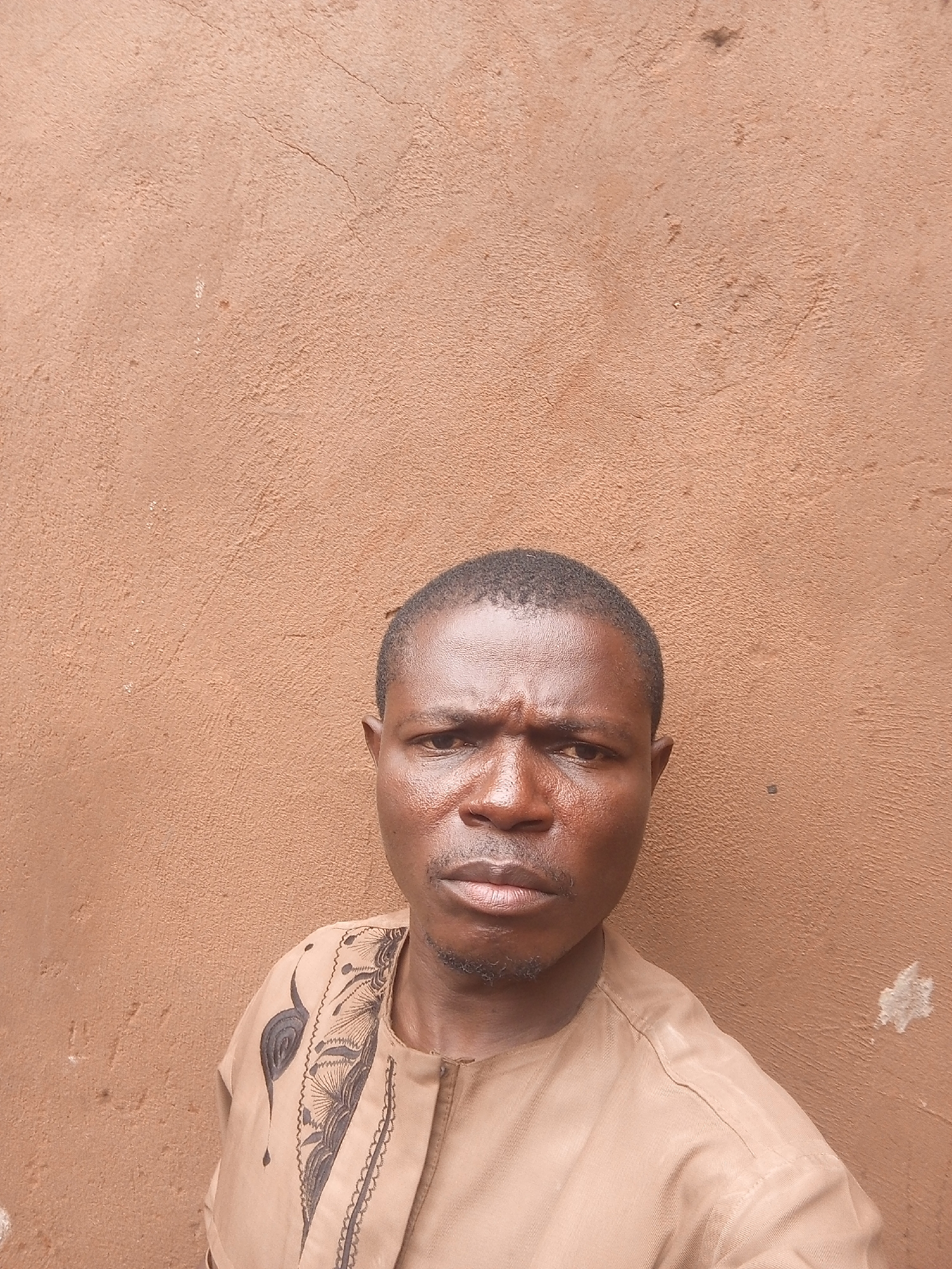 A man is standing in front of a brown wall. The man is wearing a light brown shirt with dark brown embroidery. The background appears to be outdoors, possibly in Tempure, Nigeria.