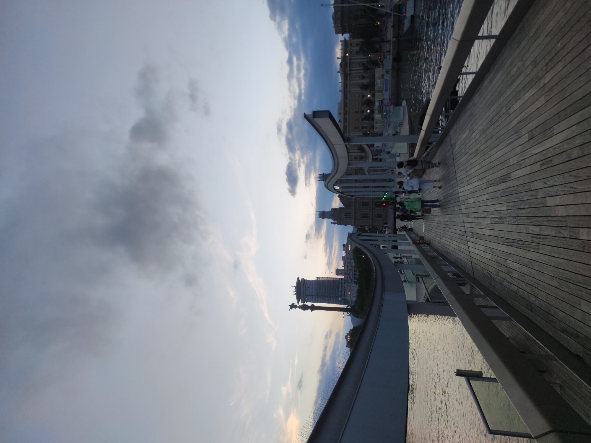 A wooden boardwalk curves away from the viewer, bordered by a metal railing. Several people walk along the boardwalk. The boardwalk is set over water, with a boat visible in the distance on the right. In the background, a city skyline features modern buildings and older architectural structures. The sky is overcast with scattered clouds. The scene appears to be a waterfront area in a city. This image is identified as being taken in Sant Boi de Llobregat, Spain.