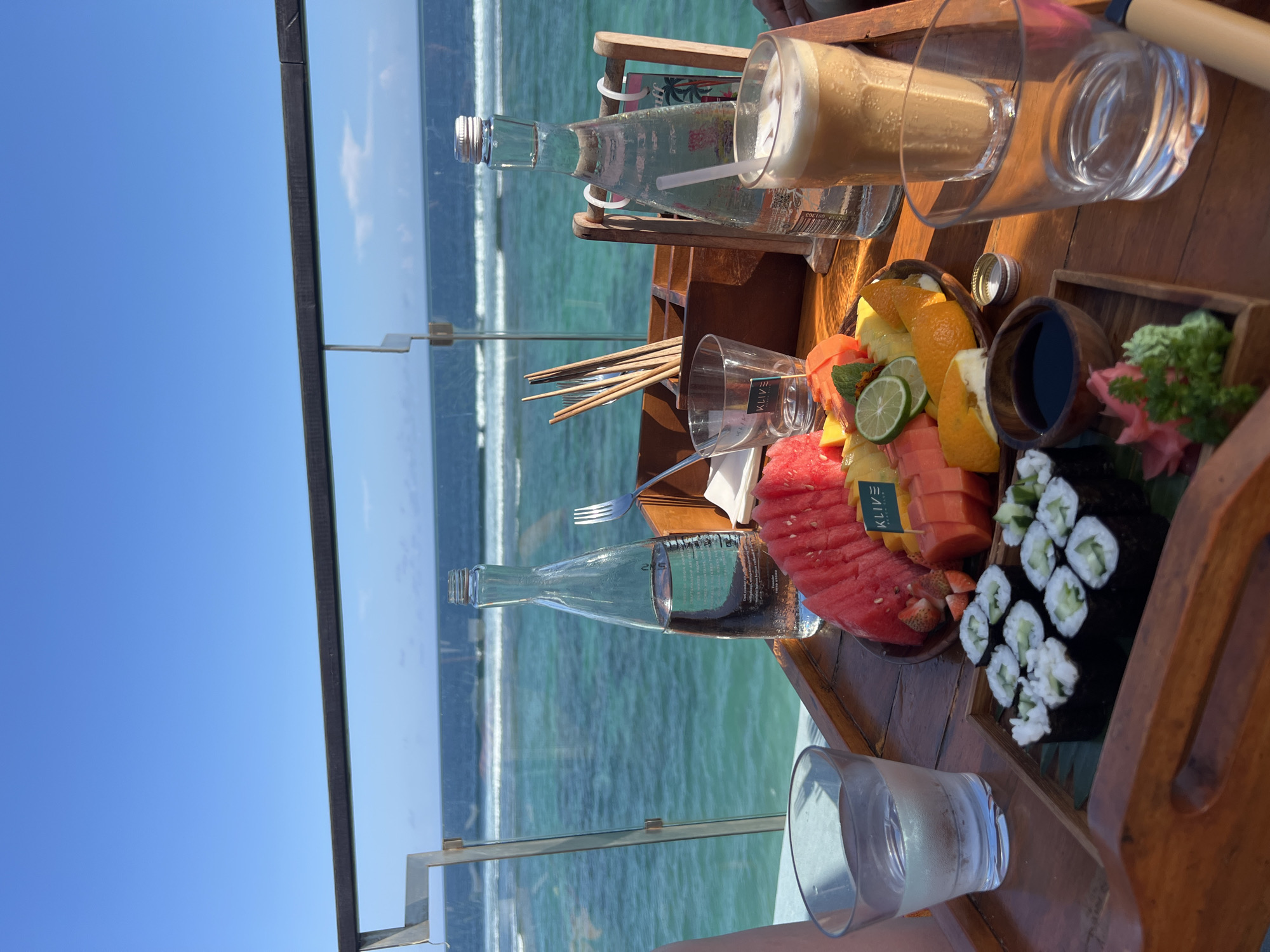 Here is a description of the image:
The image depicts a table setting with various food and beverage items, positioned against a scenic background of a blue sky and ocean. On the table, there are bottles of water, glasses, a smoothie, a wooden tray containing a fruit platter (with watermelon, papaya, orange, and lime), sushi, soy sauce, and pickled ginger. Chopsticks and a fork are also visible. The table is set against a backdrop of a clear blue sky and a turquoise ocean with visible waves. The scene evokes a sense of dining with a view, possibly in a coastal or island setting. The location and city are unknown.