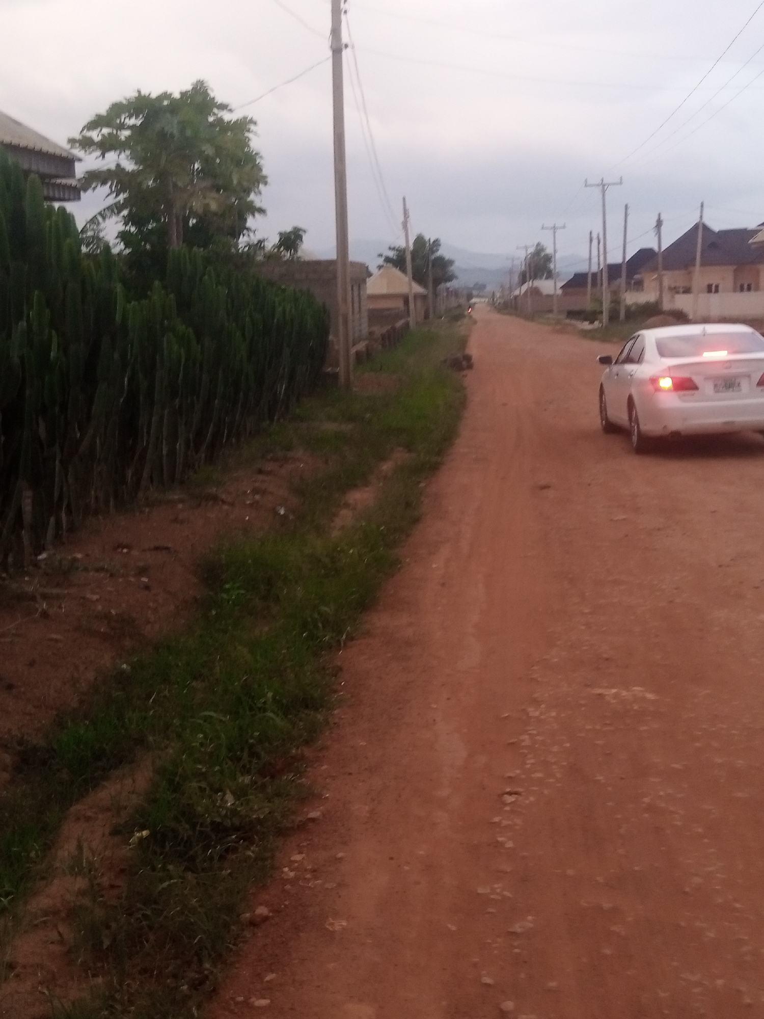 The image shows a dirt road in an urban area, possibly in Nigeria. A white car is driving down the road, and there are houses and trees on either side. The sky is overcast.