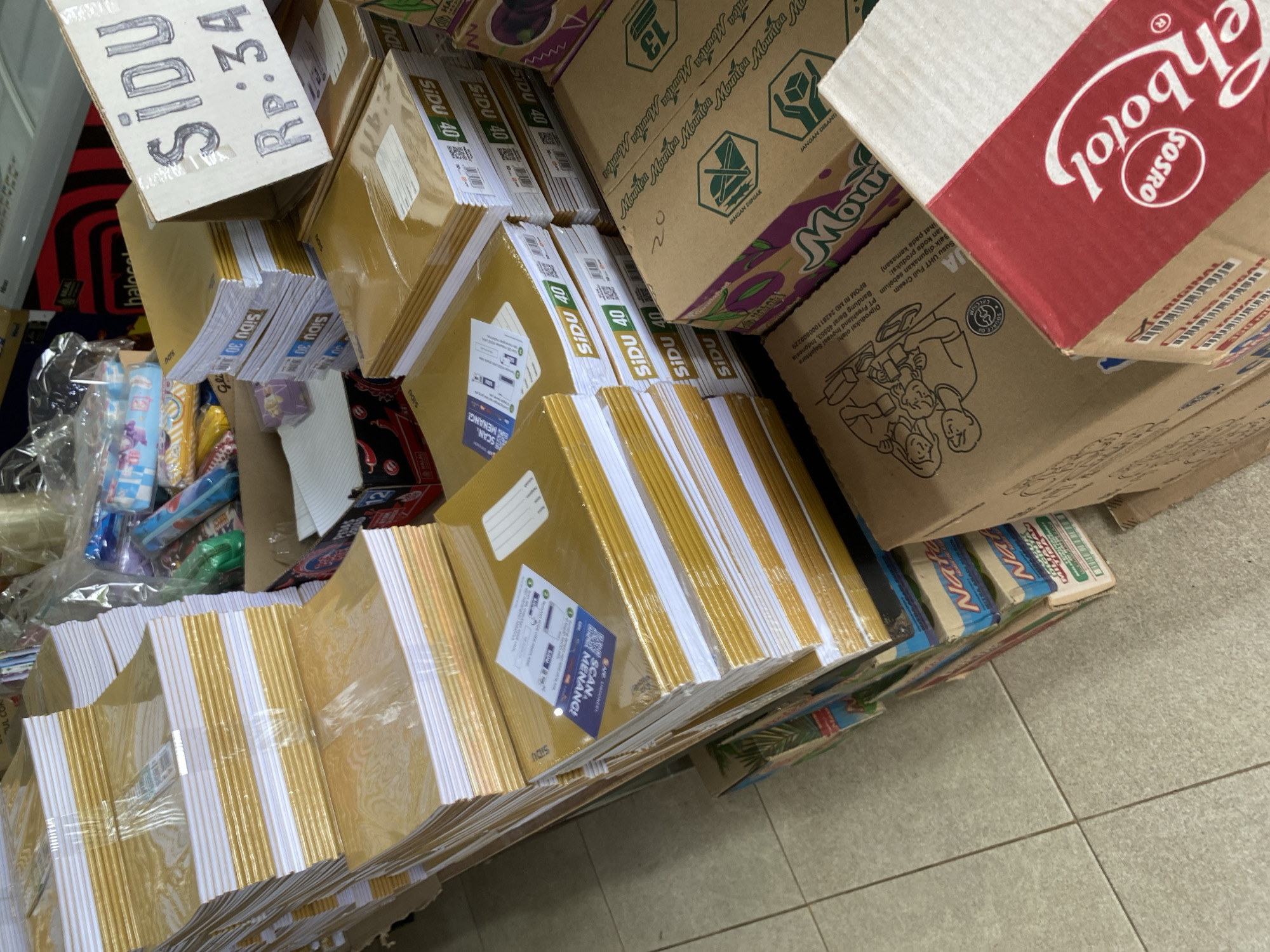The image shows a retail display of stationery and boxed food products in what appears to be a store. In the foreground, there are stacks of golden-covered notebooks with white pages, some wrapped in plastic. A price tag on a cardboard box in the upper right corner reads "SIDU Rp: 34". To the right of the notebooks, there are boxes of what seem to be food items, including one with the brand name "Mountea" visible. Further in the background, there are more stationery items and some packaged goods, possibly school supplies. The floor is tiled. The scene is consistent with a small retail shop or a market stall. The location is identified as Tanjong, Indonesia, based on the provided context. There are no people visible in the image.