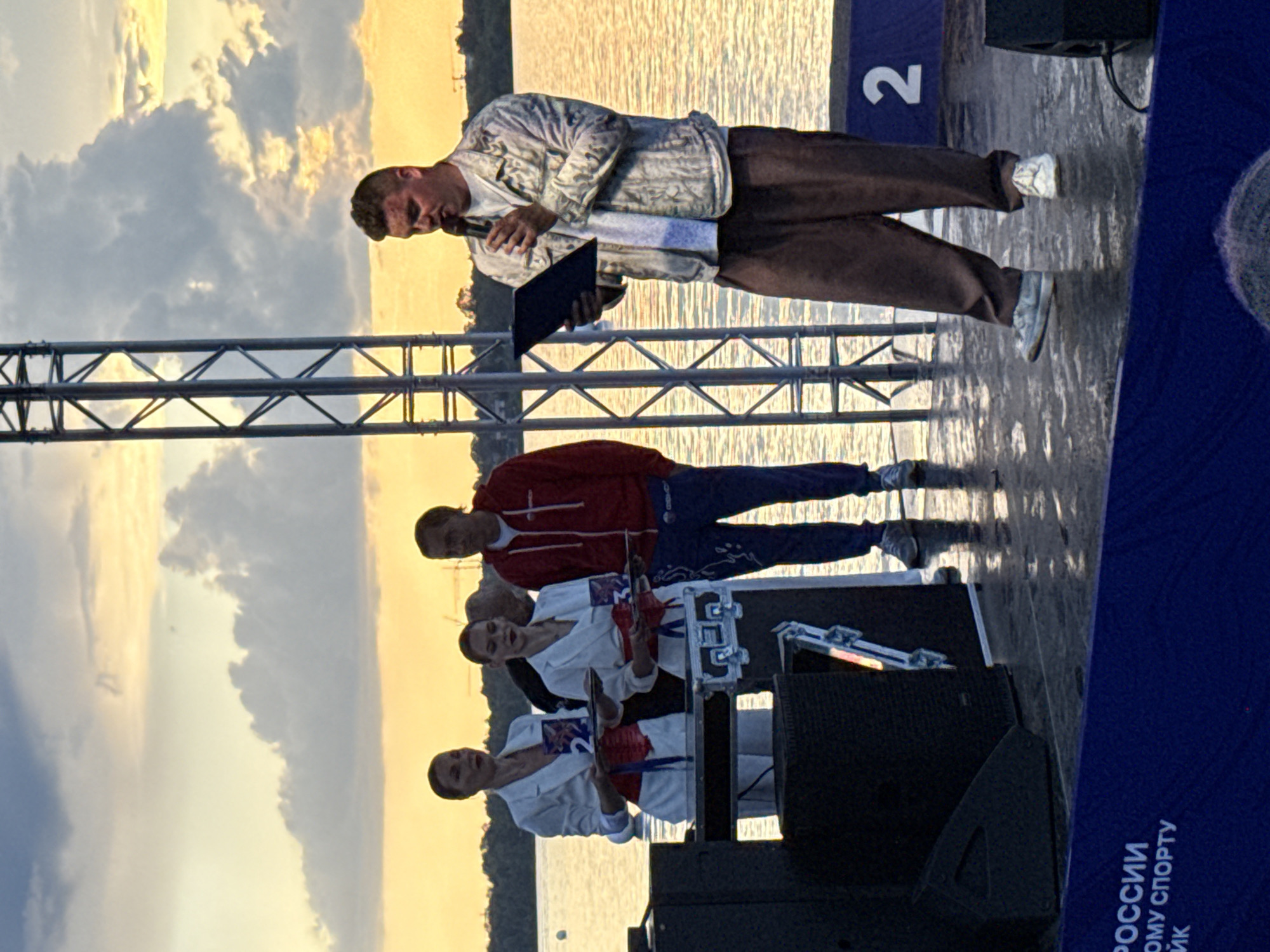 The image depicts an outdoor event at sunset, likely on a body of water, with a stage in the foreground. A man in a light-colored patterned jacket and brown pants is speaking into a microphone, holding a dark tablet or folder. He is standing on a stage with other individuals. To his left, two women in white blazers and red sashes are holding awards. Behind them, a man in a red hooded jacket and blue pants is visible. The background shows a body of water with trees on the distant shore and a dramatic sky with clouds illuminated by the setting sun. A large metal structure, possibly part of a stage or lighting rig, is present on the left side of the image. A number "2" is visible on a blue platform on the right side of the stage. Text in Russian, including "РОССИИ" and "ОМУ СПОРТУ", is visible on a blue banner at the bottom of the image, suggesting the event is related to Russian sports. The presence of the text "РОССИИ" and a sporting event context strongly suggests the location could be Perm, Russia, given the prompt's suggestion, though visual evidence within the image alone does not confirm this specific city. The overall scene appears to be an awards ceremony or presentation at a sporting competition.