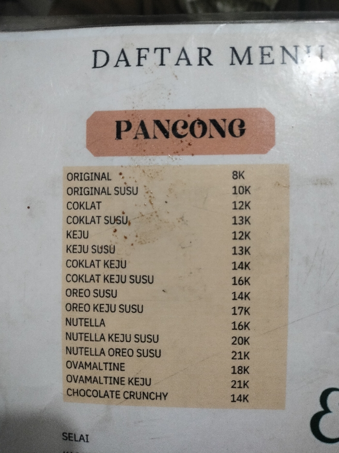 The image displays a close-up of a laminated food menu, likely from an eatery in Dramaga, Indonesia. The menu appears worn, with visible dust and specks scattered across its surface. At the top, partially cropped, the text "DAFTAR MENU" (Menu List) is visible.
The central part of the menu is dedicated to a specialty item called "PANCONG," printed in a large, decorative font on a reddish-brown banner. Below this heading, a list of various "Pancong" flavors and their prices is presented. These include "ORIGINAL" (8K), "ORIGINAL SUSU" (10K), "COKLAT" (12K), "COKLAT SUSU" (13K), "KEJU" (12K), "KEJU SUSU" (13K), "COKLAT KEJU" (14K), "COKLAT KEJU SUSU" (16K), "OREO SUSU" (14K), "OREO KEJU SUSU" (17K), "NUTELLA" (16K), "NUTELLA KEJU SUSU" (20K), "NUTELLA OREO SUSU" (21K), "OVAMALTINE" (18K), "OVAMALTINE KEJU" (21K), and "CHOCOLATE CRUNCHY" (14K). The "K" suffix indicates prices in thousands of Indonesian Rupiah. A subsequent menu section, starting with "SELAI" (Jam), is partially visible at the bottom left. No individuals or specific activities are depicted; the image focuses solely on the menu as an object.