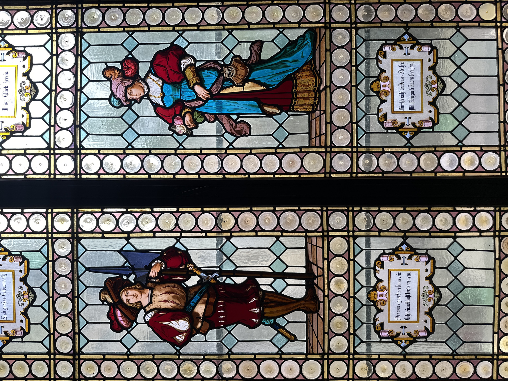 The image shows two stained glass panels, one above the other. The top panel depicts a couple, a man in a red jacket and a woman in a blue dress, embraced. The man is holding flowers. Next to them is a panel with text in an ornate frame. The bottom panel shows a man in armor holding a sword and a banner, with another panel next to him containing text in a similarly styled frame. The stained glass panels are framed by borders of circular glass pieces.