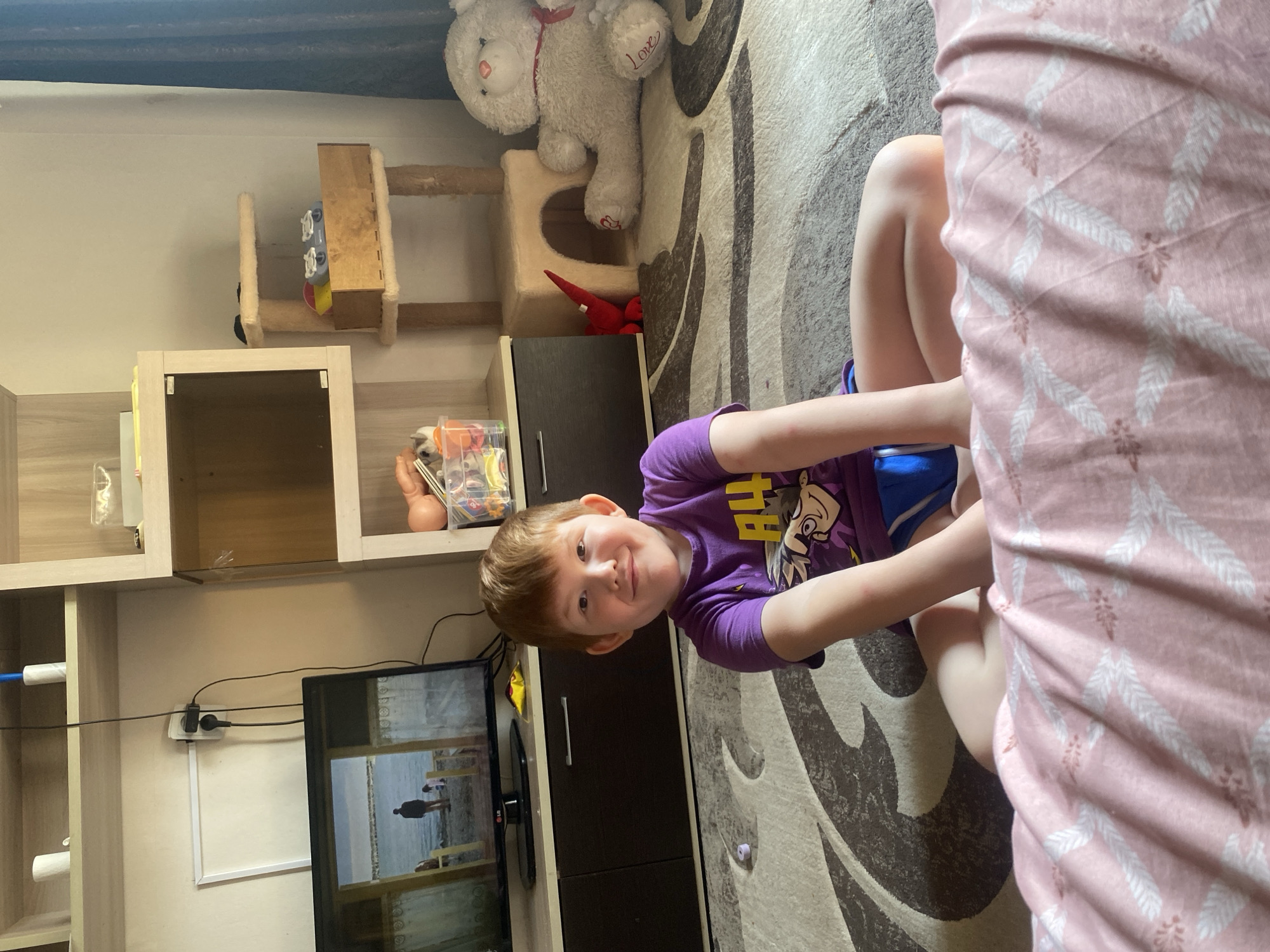 A young boy with reddish-brown hair smiles at the camera. He is wearing a purple t-shirt with a cartoon character and blue shorts. He is sitting on a rug with a patterned design. Next to him, a pink blanket with a white leaf pattern is draped over furniture. In the background, there is a shelf unit with various items, including a white teddy bear and a red dinosaur toy. A television screen shows a scene of people by the water. Electrical outlets and cords are visible on the wall behind the TV. The scene appears to be indoors, in a home. The lighting suggests it is daytime.