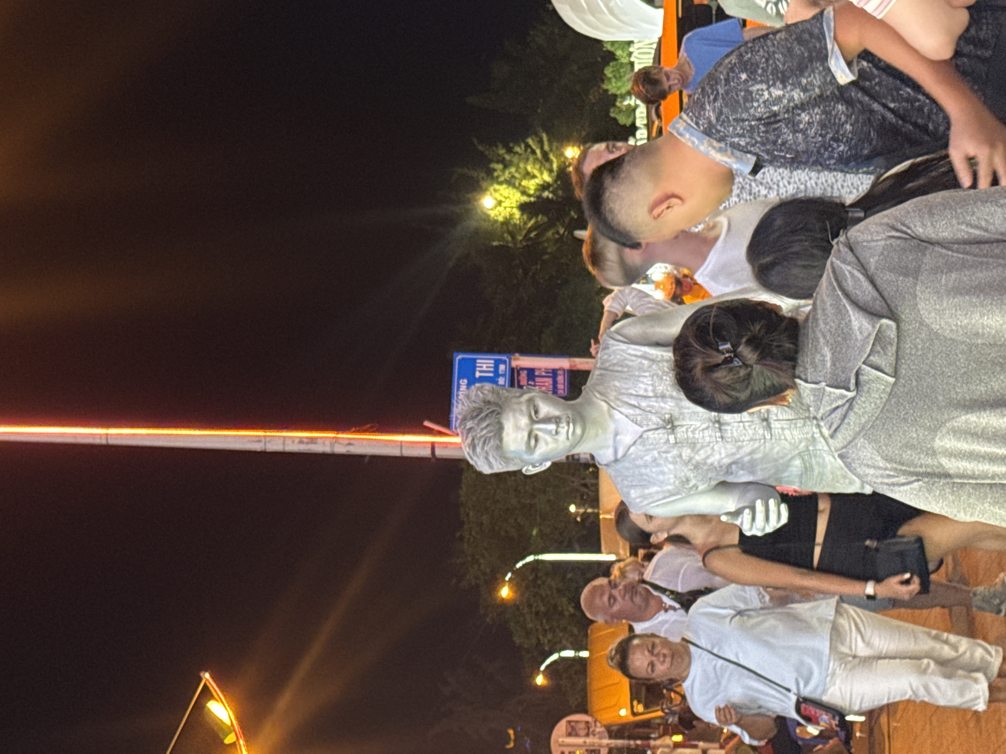 The image depicts a street scene at night in Nha Trang Ward, Vietnam, featuring a live statue performer painted entirely in silver. The performer stands centrally, facing forward with a neutral expression. Surrounding the performer is a crowd of onlookers, some of whom are looking at the performer, while others are engaged in conversation or looking away. The environment includes streetlights, trees, and a bus in the background. The lighting suggests an urban setting with ambient illumination from streetlights and possibly neon signs. One sign visible in the background appears to be in Vietnamese. The overall atmosphere is that of a lively street or public gathering at night.